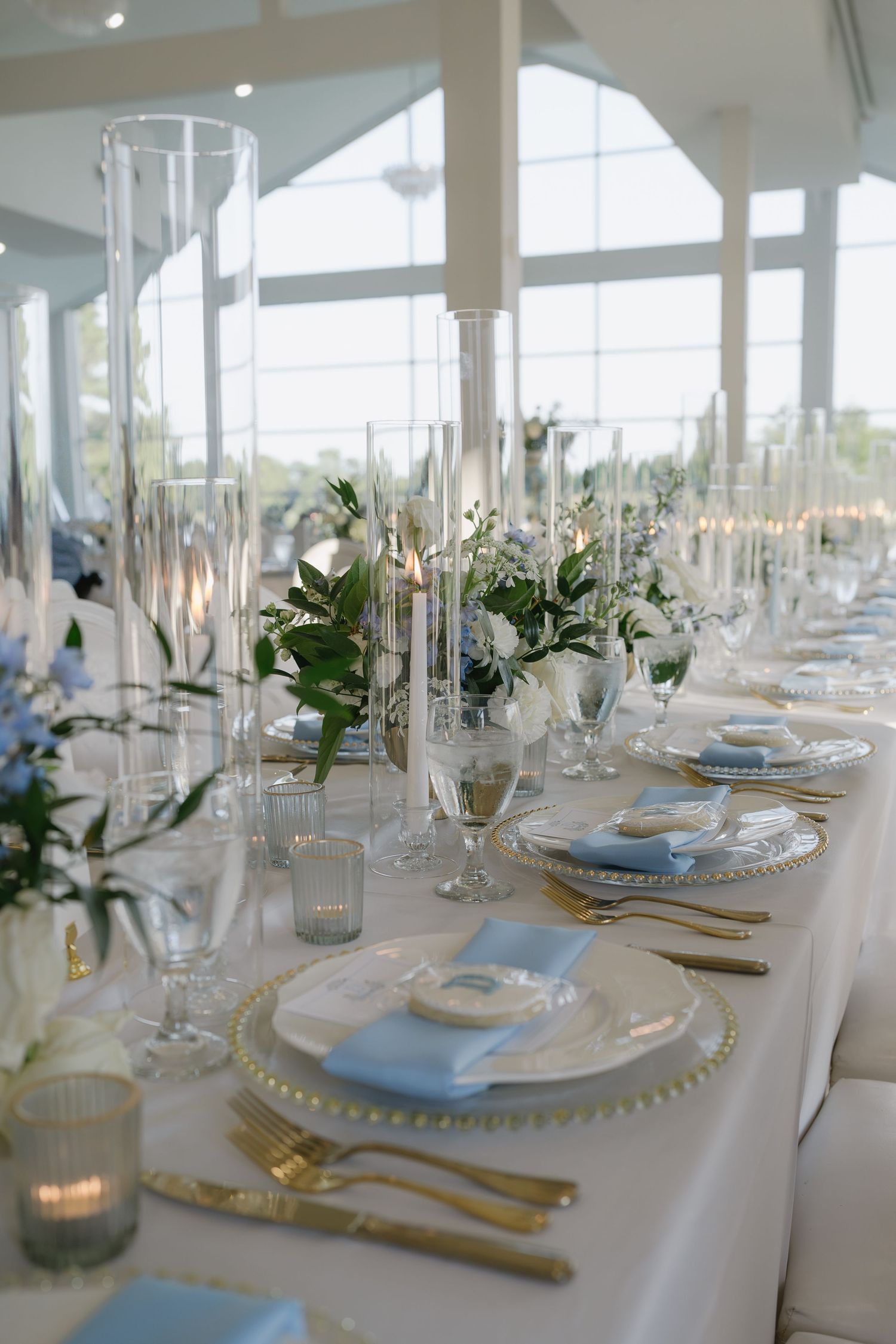 Elegant wedding reception table with gold cutlery, glass chargers, blue napkins, tall candle holders, and white floral centerpieces.