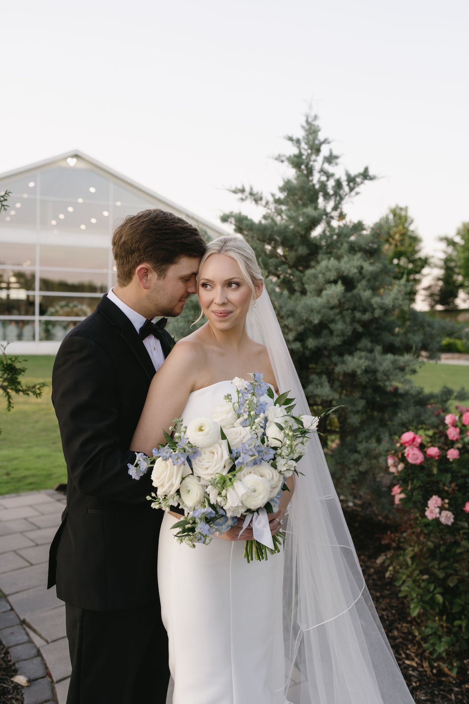Bride and groom pose outdoors, she holds white and blue bouquet, wearing strapless gown with veil, he wears black tuxedo.