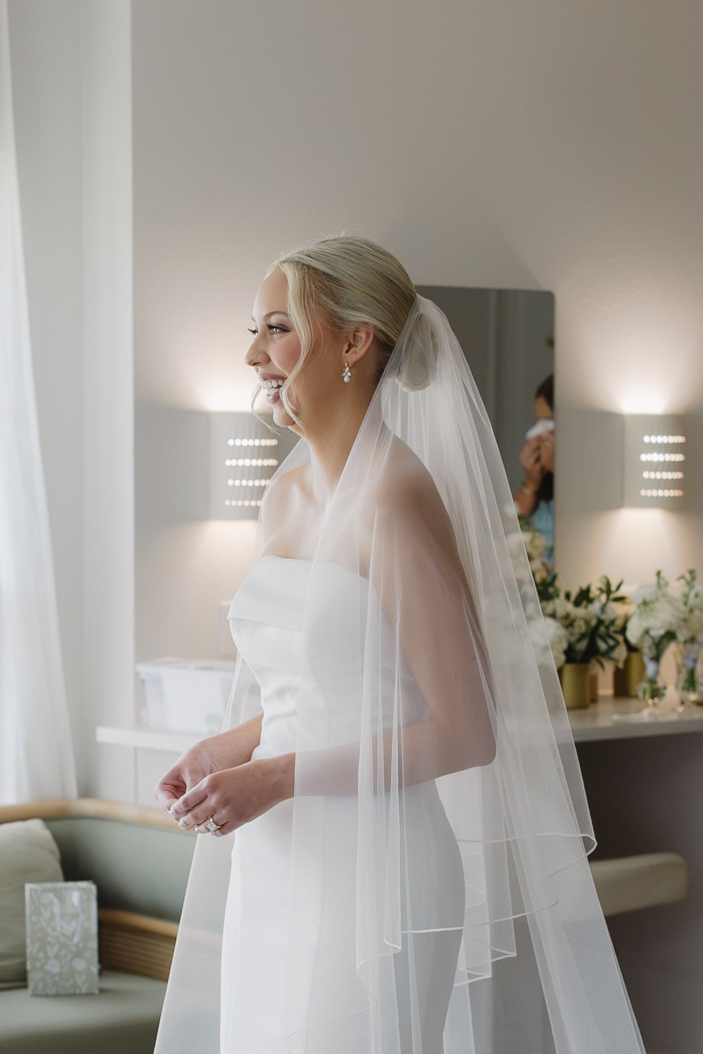 Smiling bride in a white strapless gown and layered veil stands in a softly lit bridal suite.