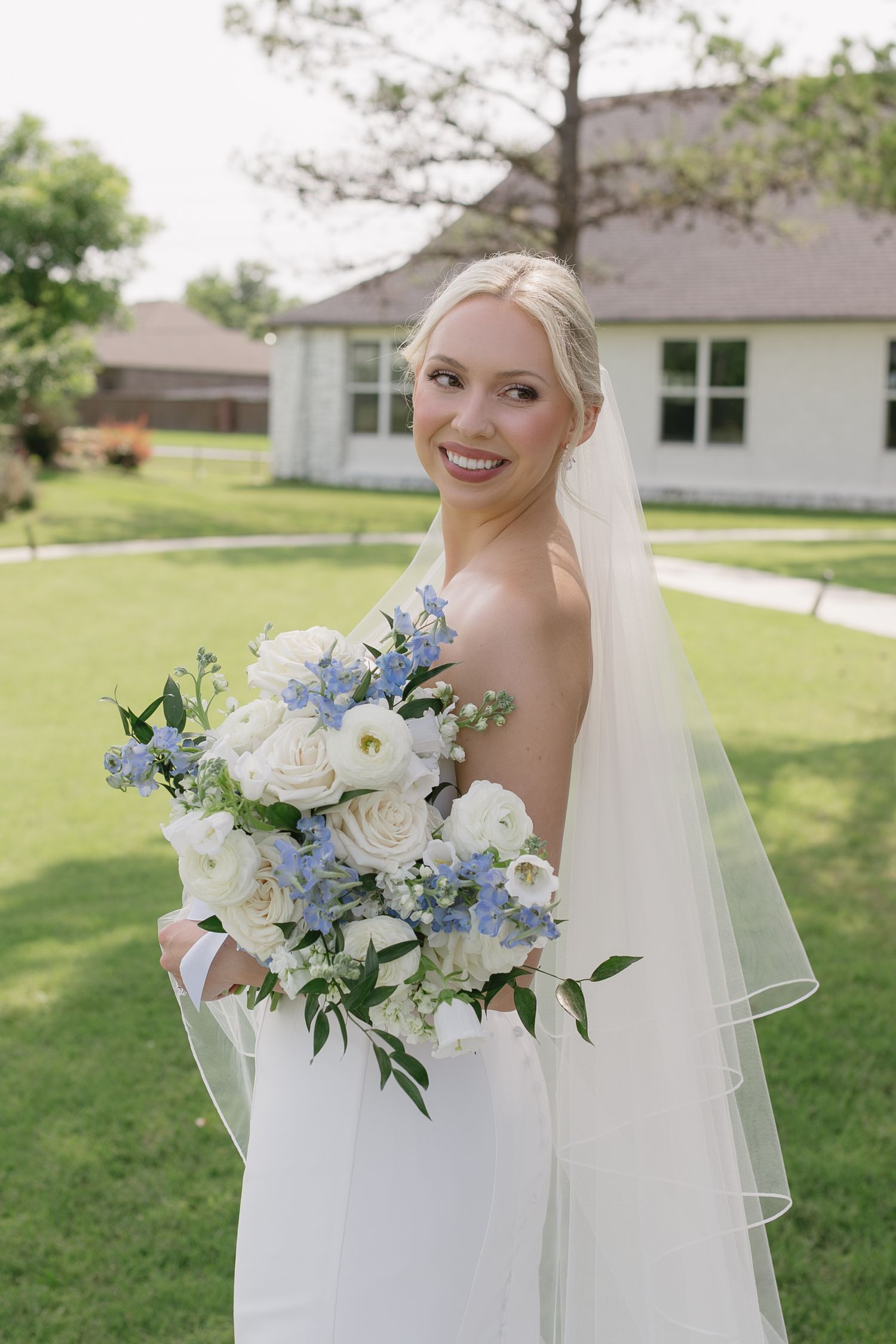 Smiling bride in white gown and veil holds blue and white floral bouquet outdoors on green lawn.
