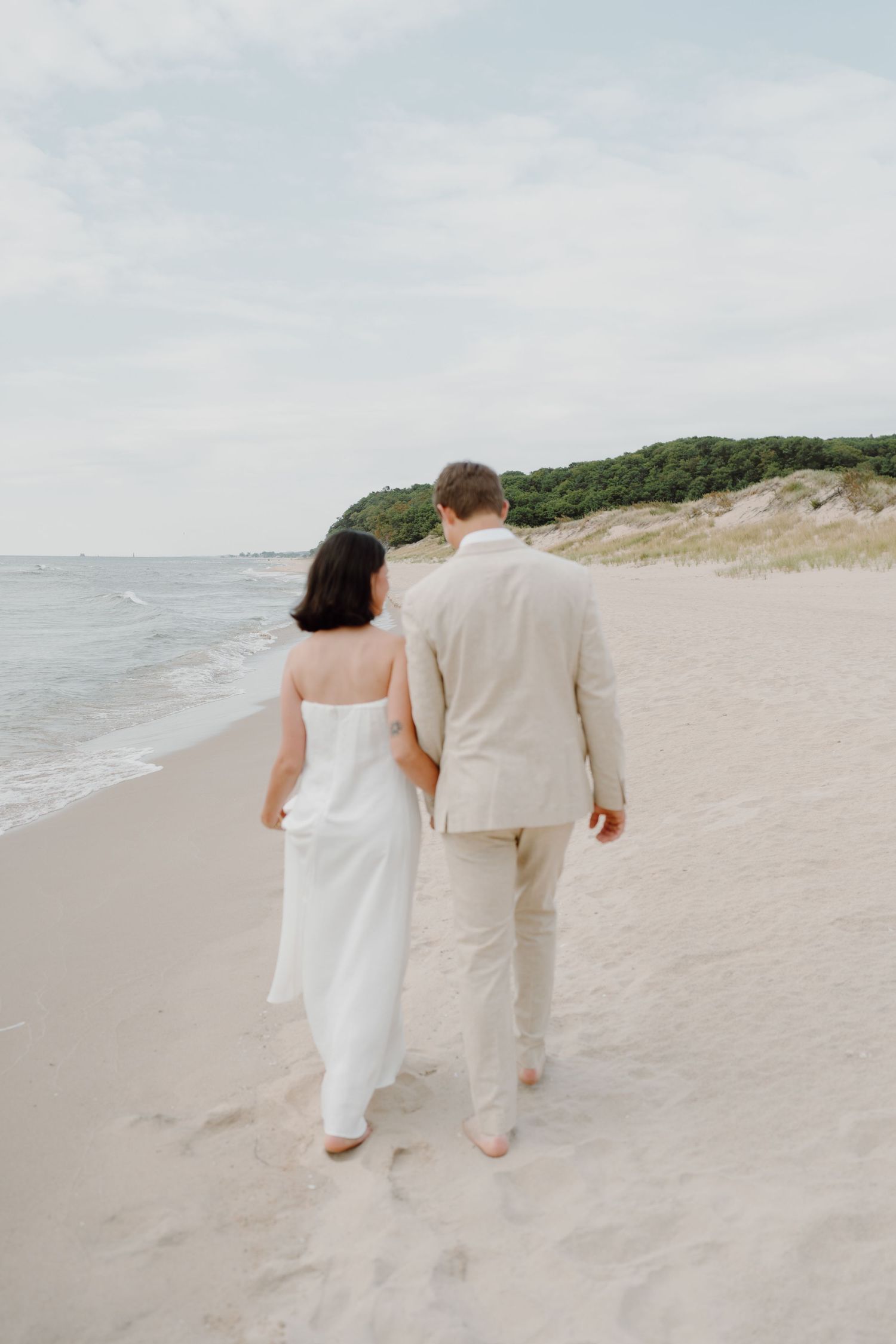 A couple enjoys a romantic beach walk against a backdrop of sand dunes and ocean waves.