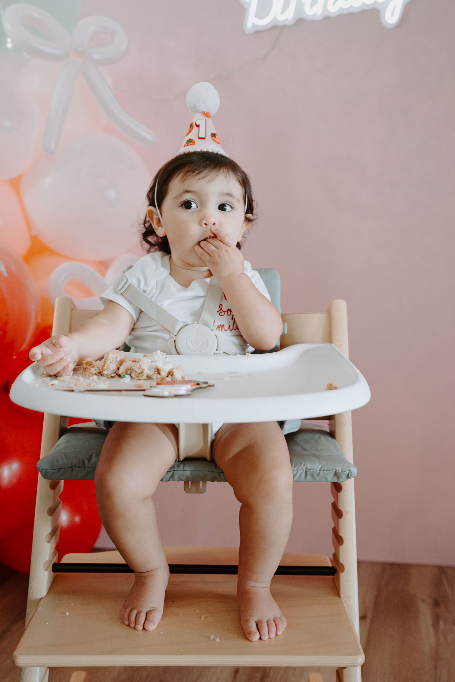 Small high chair against pink wall with 'Happy Birthday' neon sign visible.