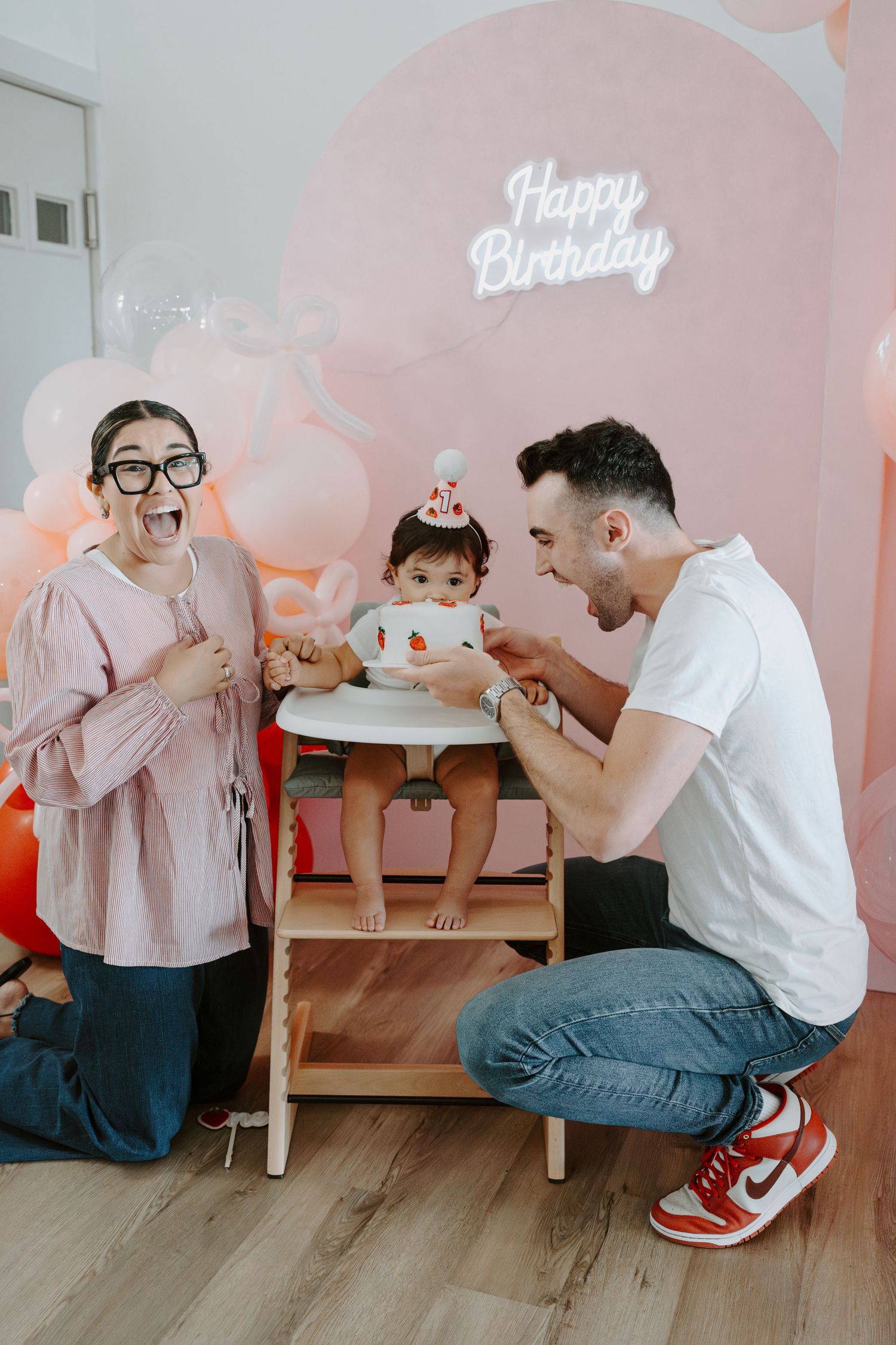 Excited family celebrating baby's birthday with animated expressions and pink party decor.