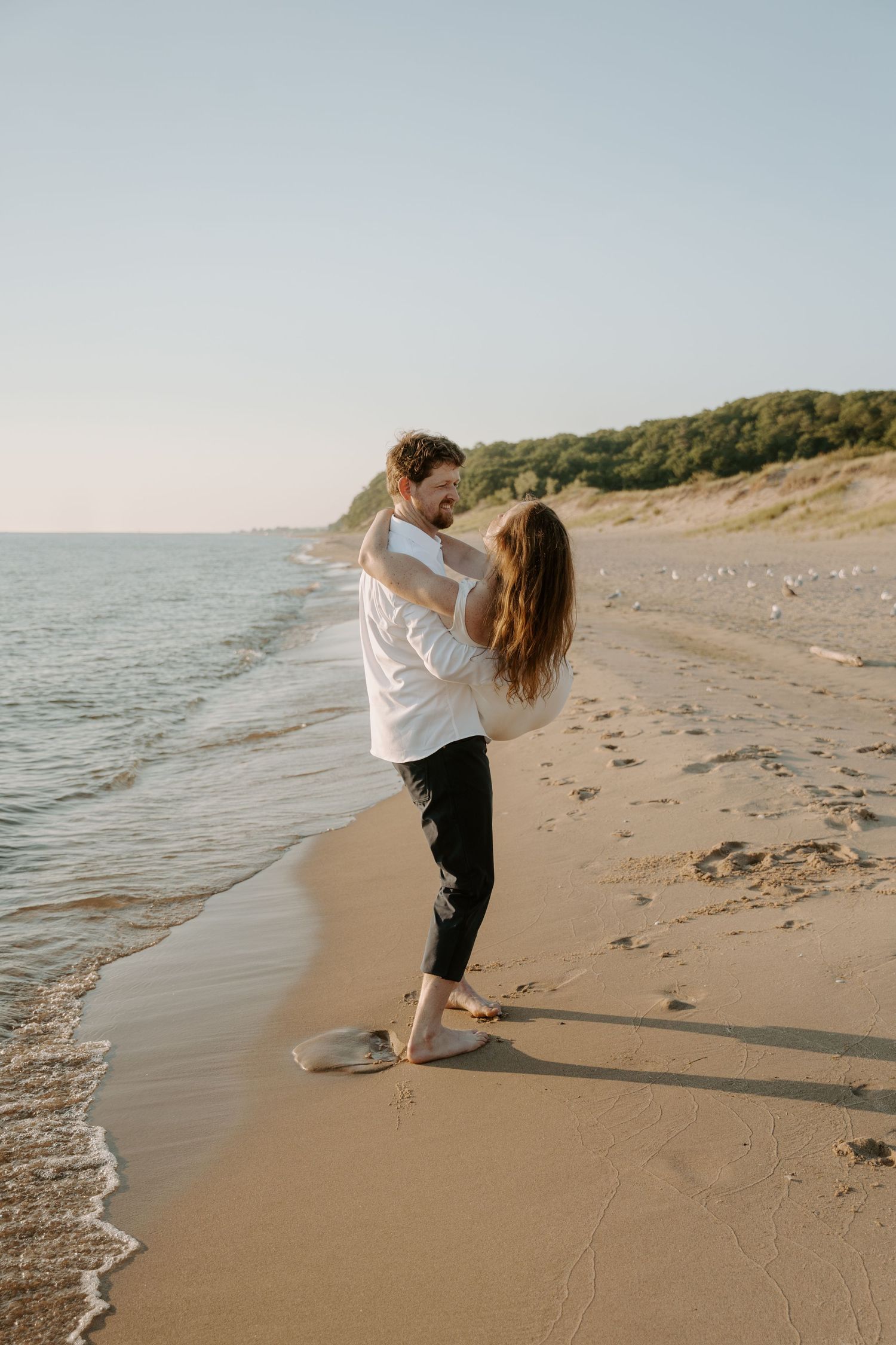 Couple shares playful lift pose along ocean shoreline at sunset.