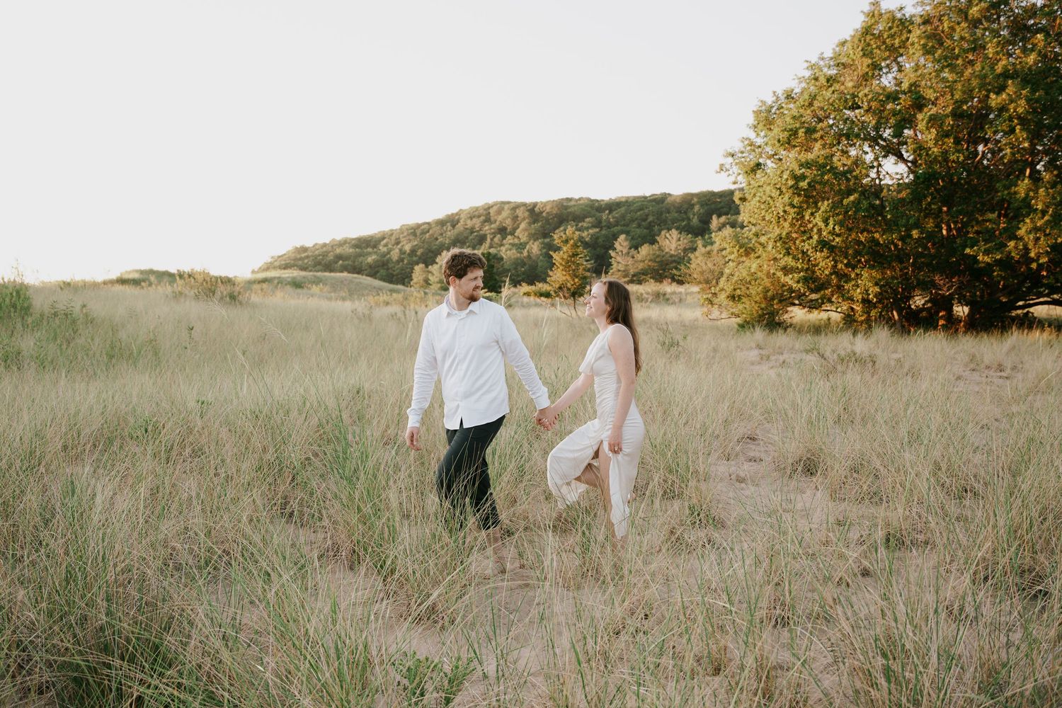 A couple strolls hand in hand through tall grass in a golden field at sunset.