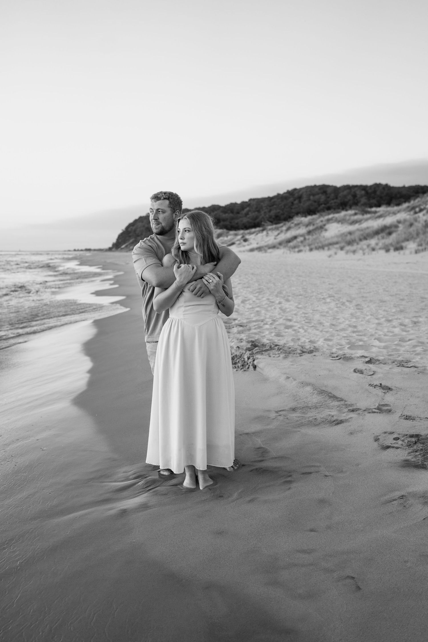 A black and white photo captures a couple's playful moment on a sandy beach at sunset.