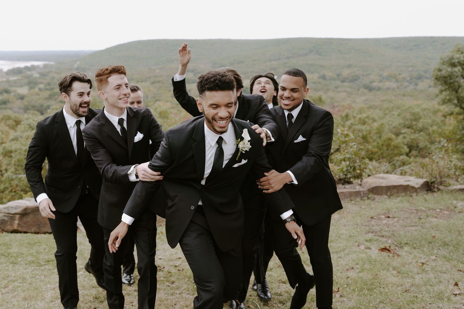 Wedding party members in black suits strike dynamic poses on a scenic overlook.
