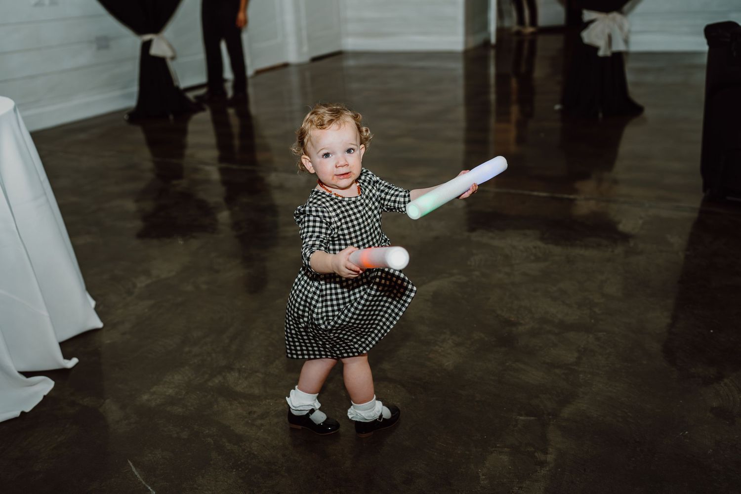 Small child in checkered dress holding light stick on dark reception dance floor.