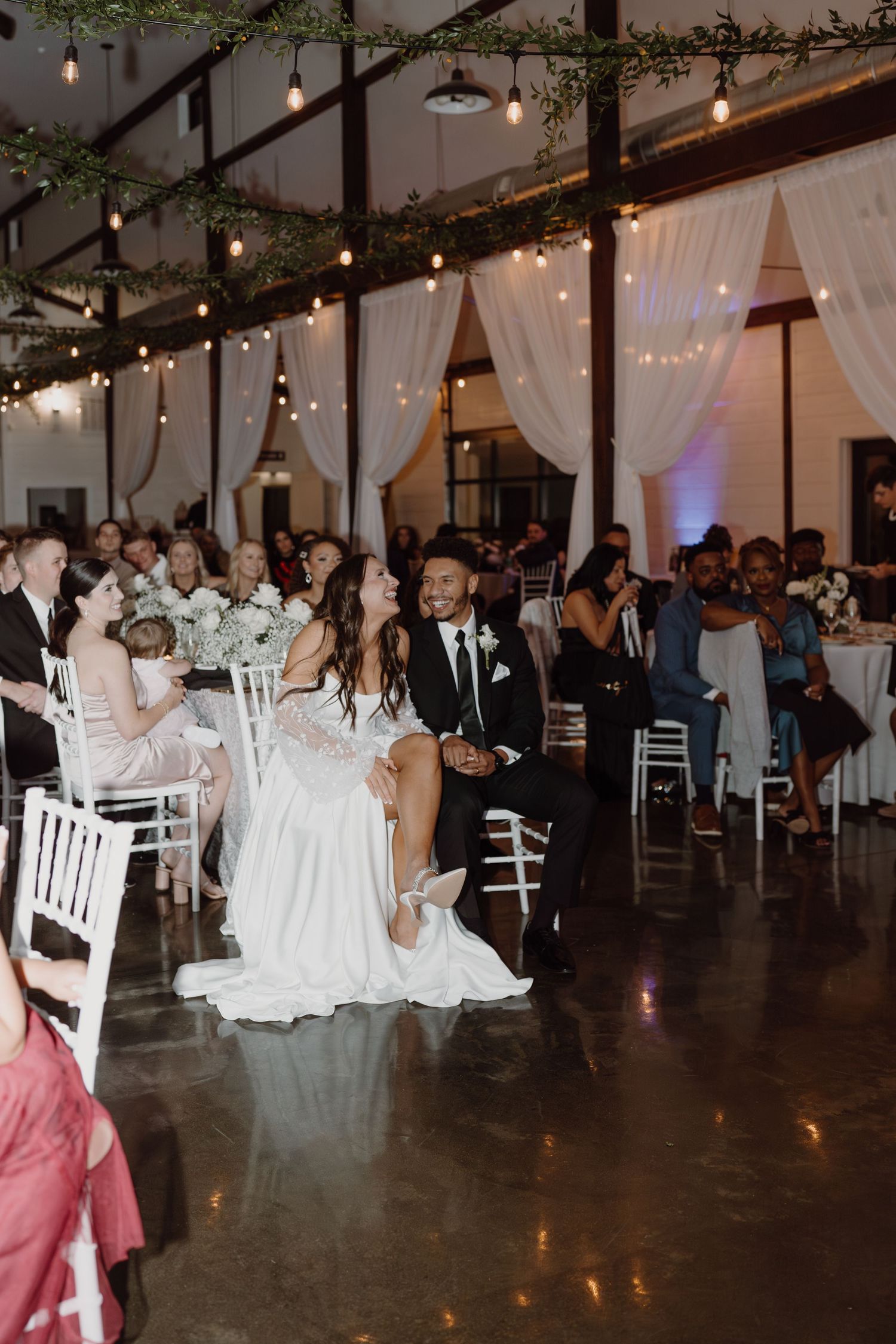 A festive wedding reception scene with string lights, greenery, and white chairs.