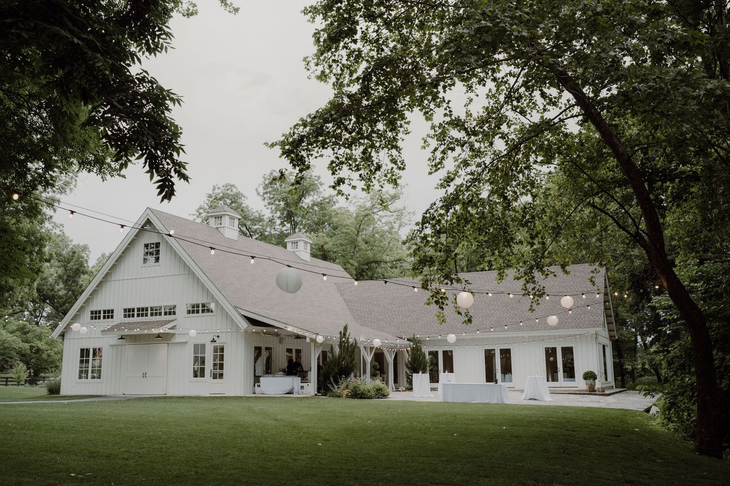 Rustic white barn wedding venue with wraparound porch surrounded by green trees.