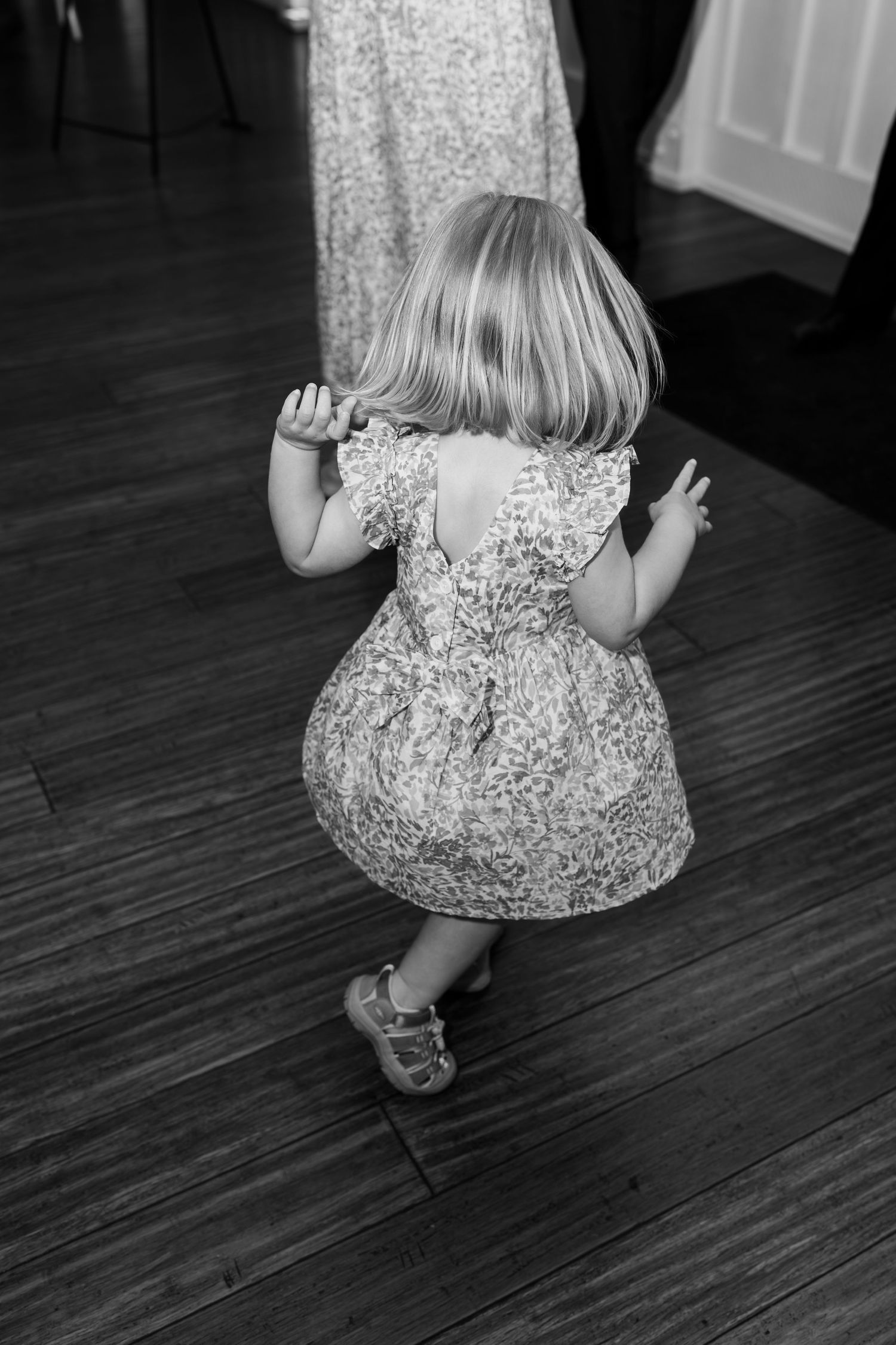 Black and white photo of a small child in a floral dress sitting on hardwood floors.