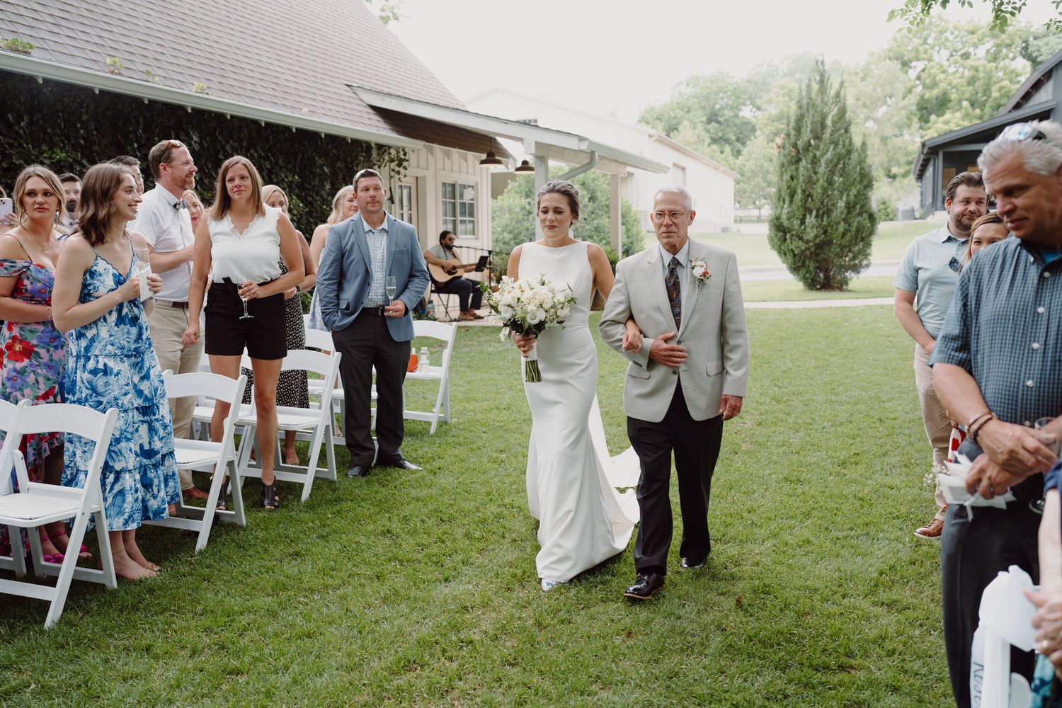 A wedding ceremony processional on a lawn with guests seated in white chairs and buildings in background.