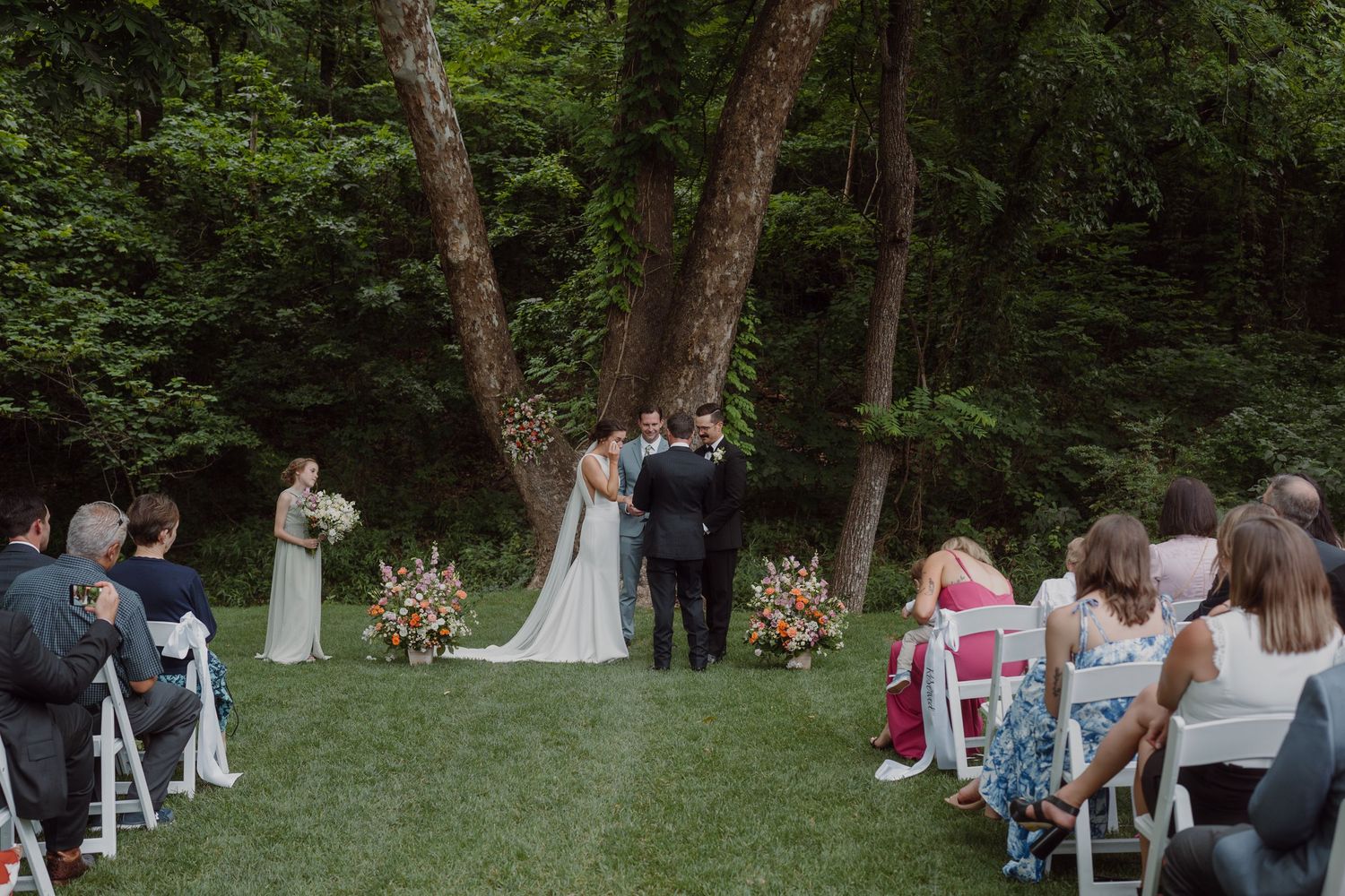 Wide view of wedding guests seated in white chairs during an outdoor forest ceremony.