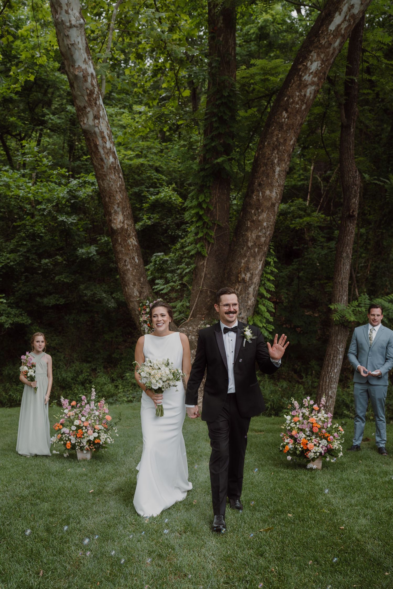 Wedding party walking through a green forest setting with colorful floral arrangements and white chairs.