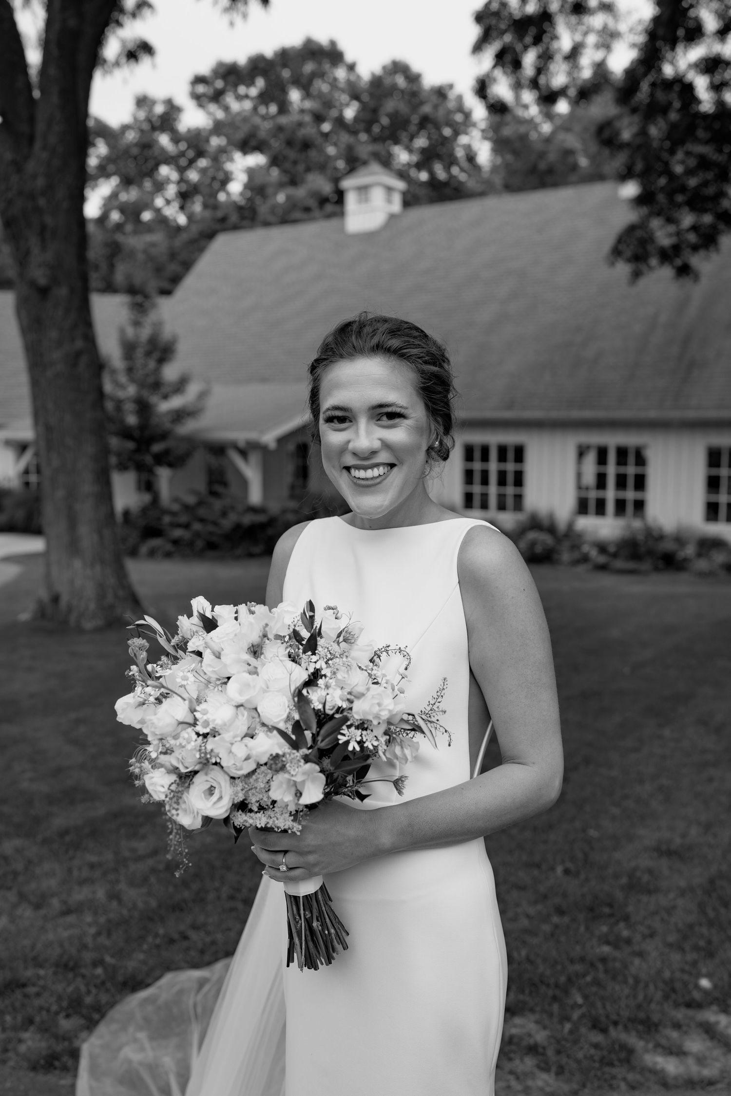 A black and white photo of a bride holding a white bouquet near a rustic building.