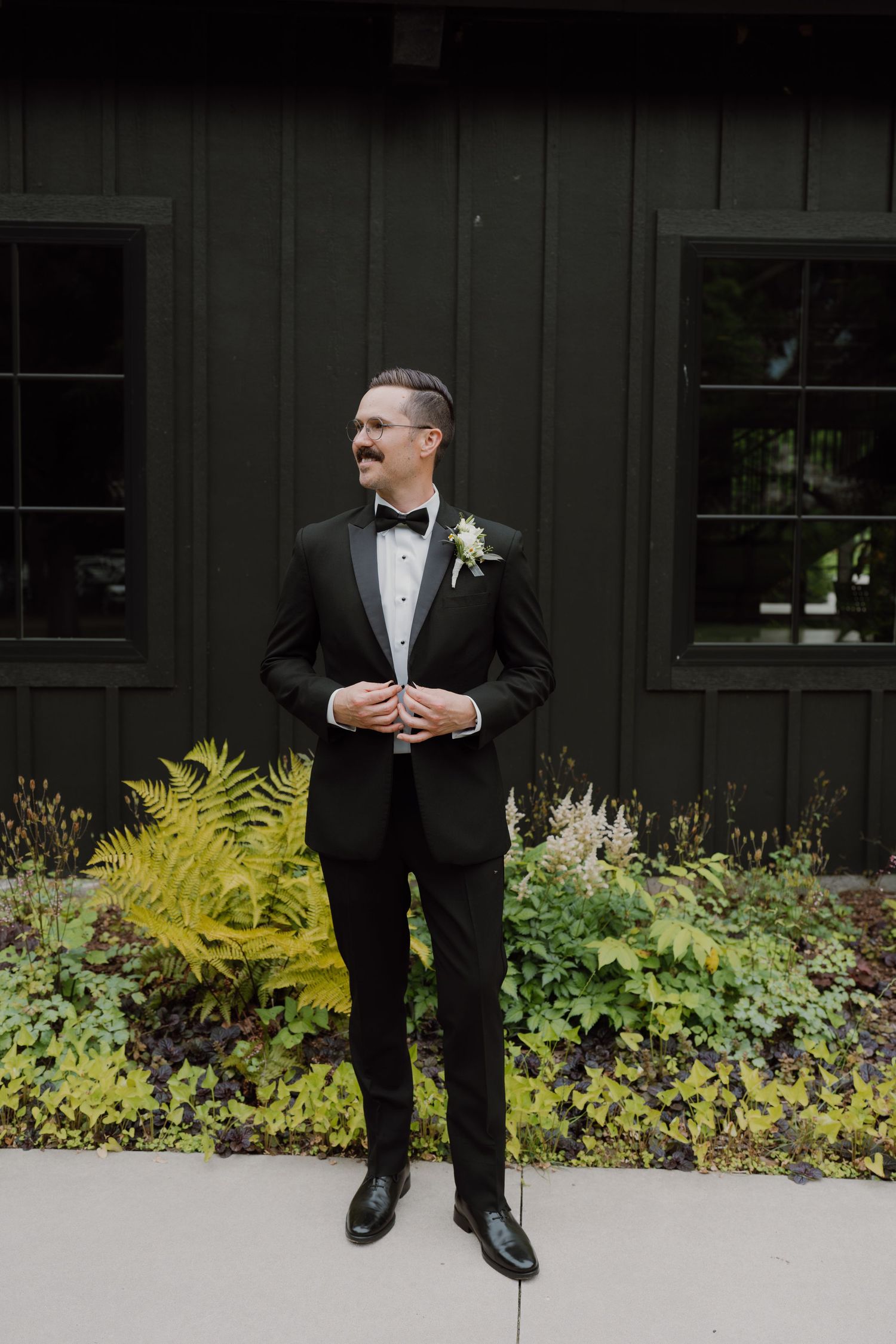 Classic groom portrait in black tuxedo with bowtie against dark wall and garden backdrop.