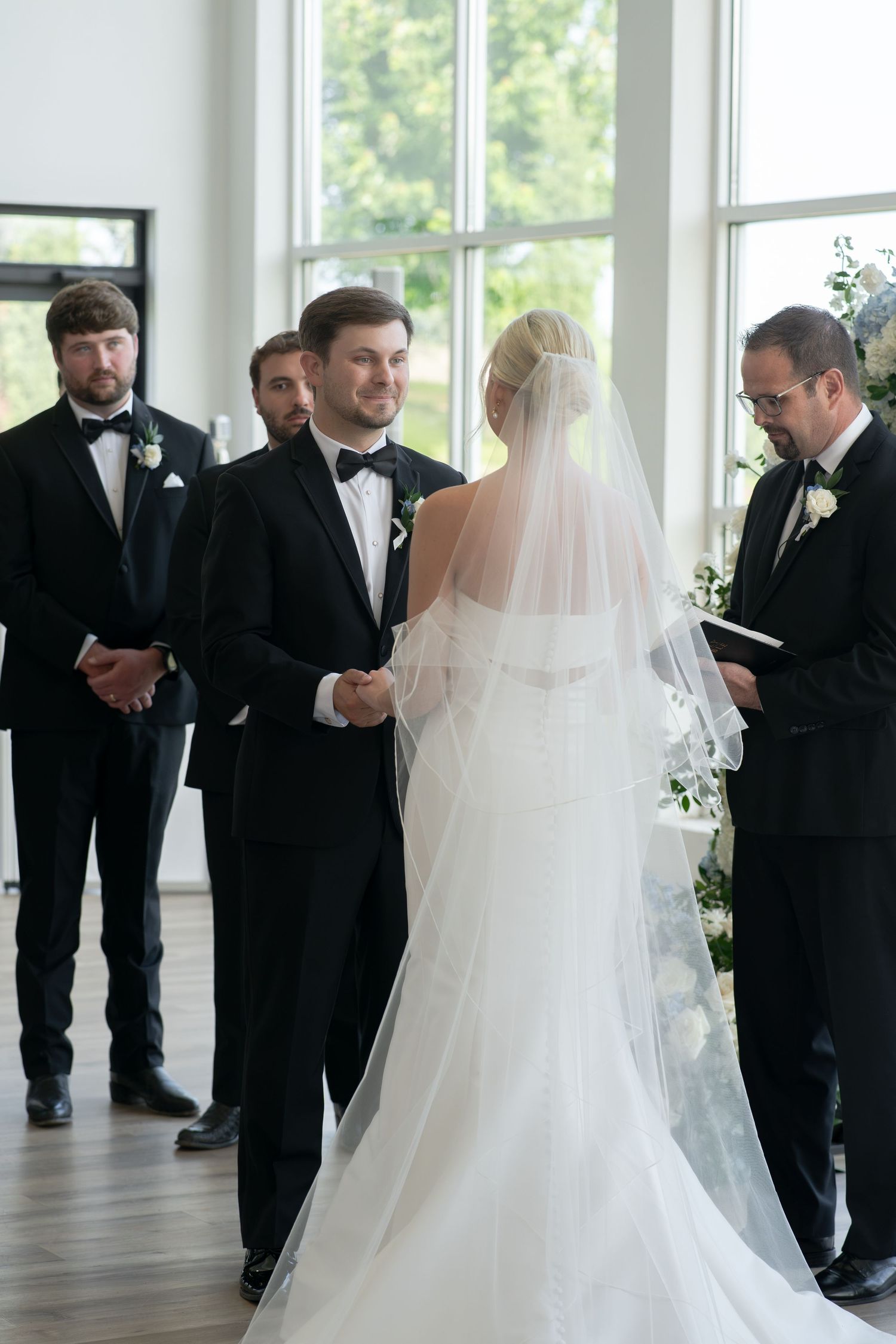 Bride and groom exchange vows at elegant indoor wedding ceremony with groomsmen and officiant in black tuxedos.
