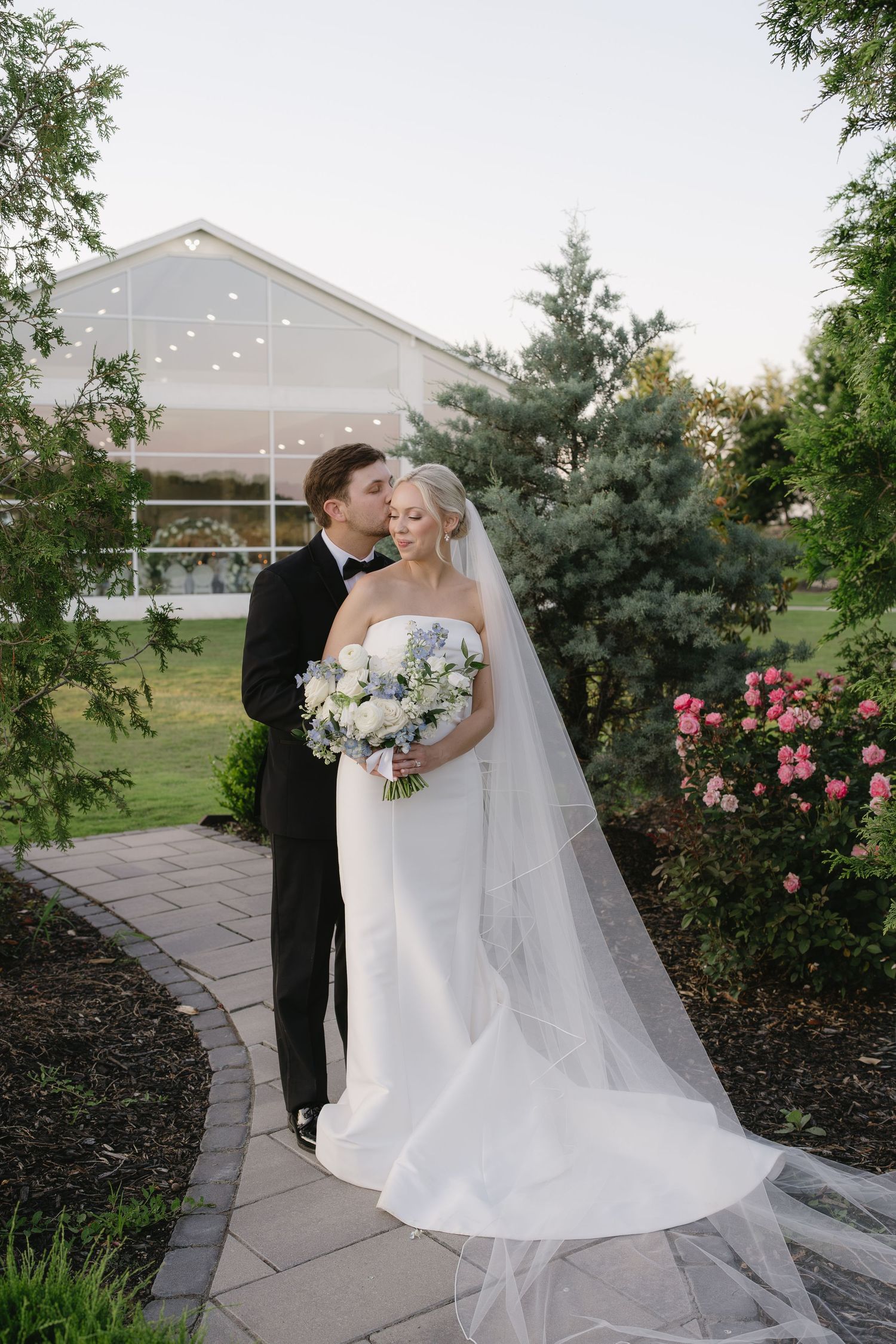 Bride and groom share a romantic moment on a garden path, bride in white gown with veil holding a bouquet.