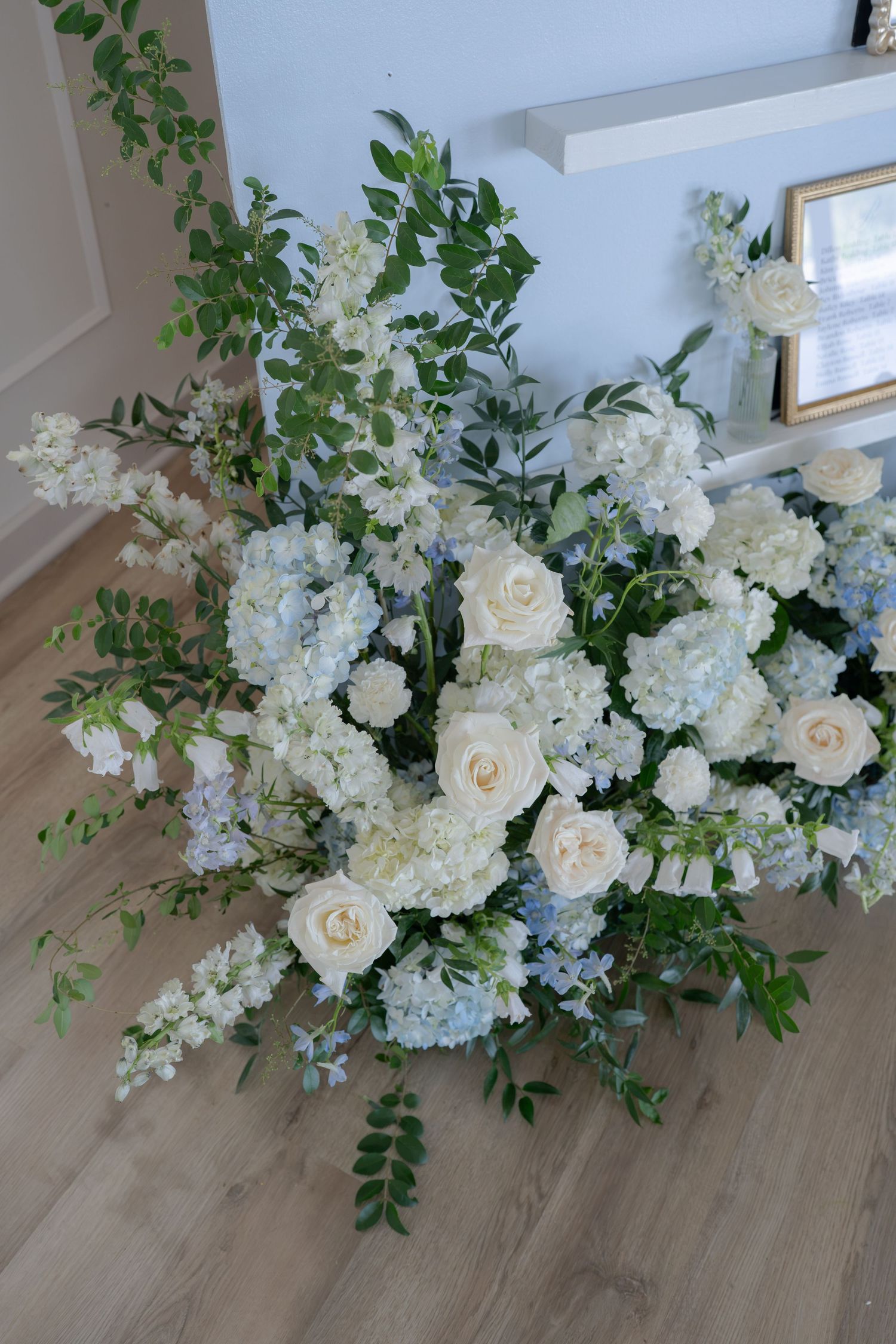 Lush white roses and blue hydrangeas floral arrangement with greenery on hardwood floor near staircase.