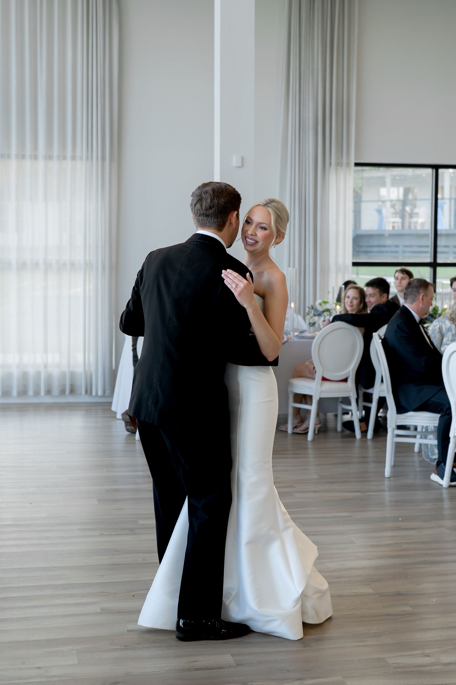 Bride and groom share their first dance in a bright modern wedding venue while guests watch from round white chairs.