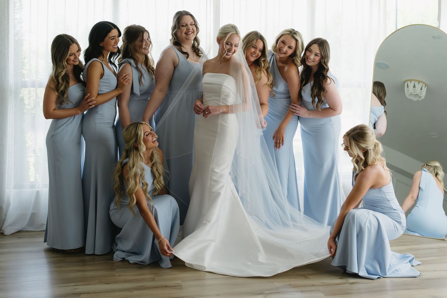 Bride in white gown surrounded by bridesmaids in light blue dresses, smiling and celebrating together indoors.