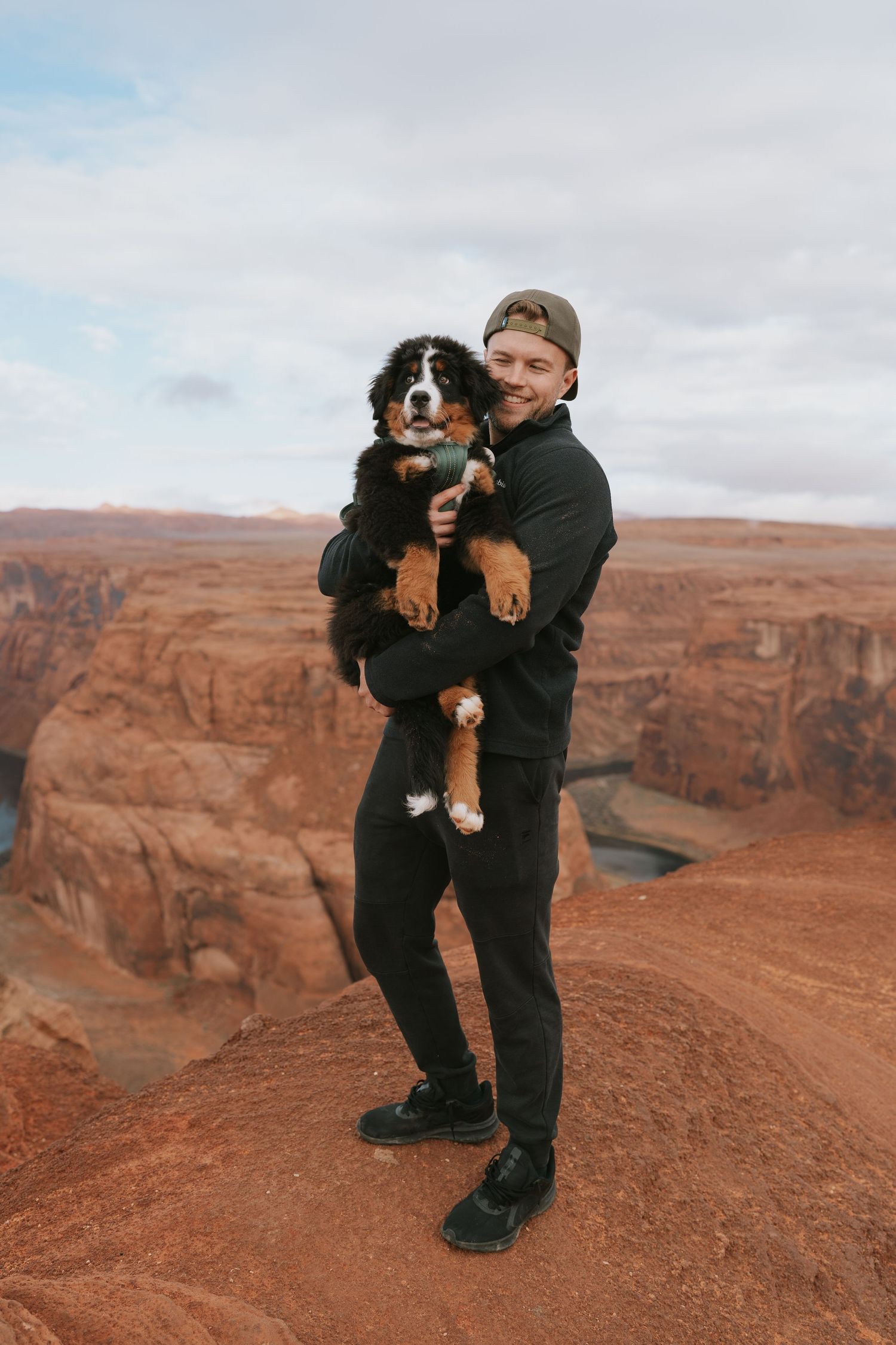 Person holds a Bernese Mountain Dog against backdrop of red desert cliffs.