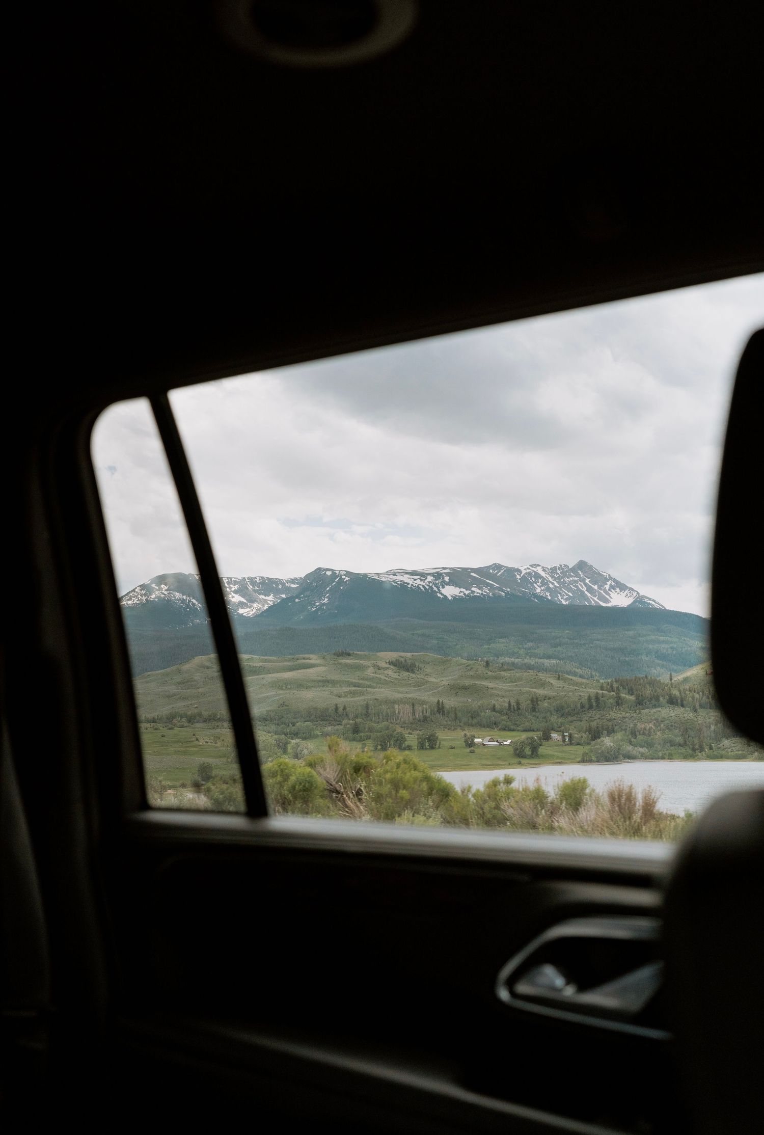 Mountain landscape viewed through car window showing snow-capped peaks and rolling hills.