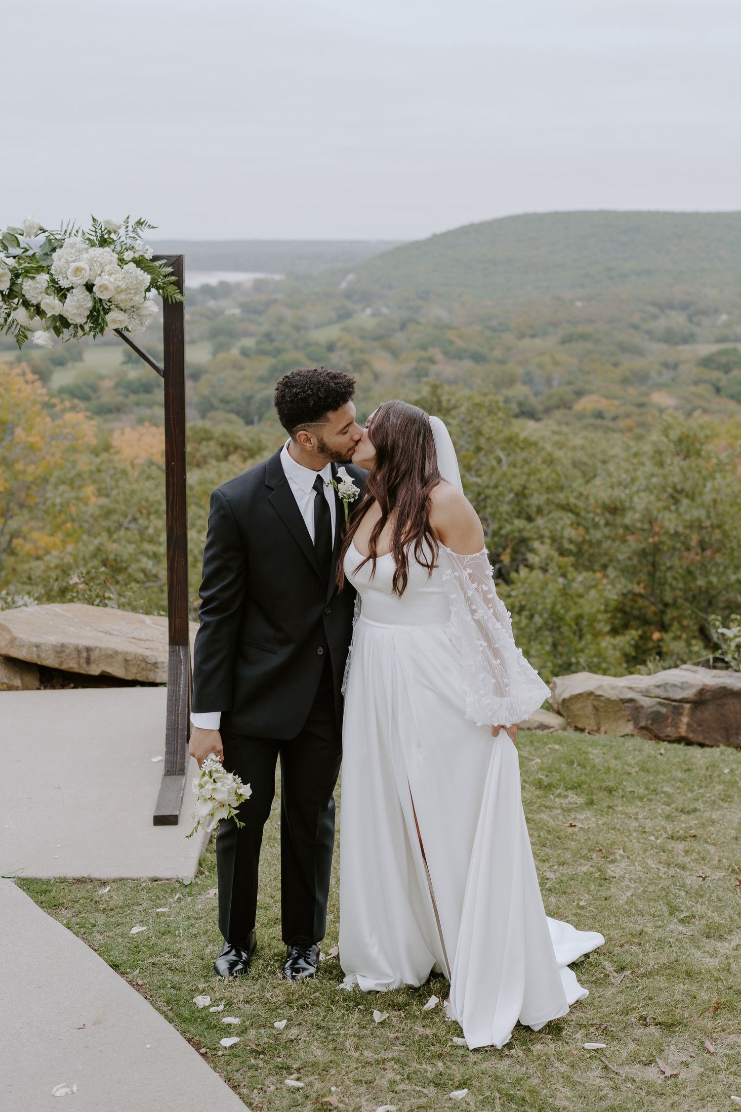 A couple in wedding attire shares tender moments along a hilltop path with mountain views.
