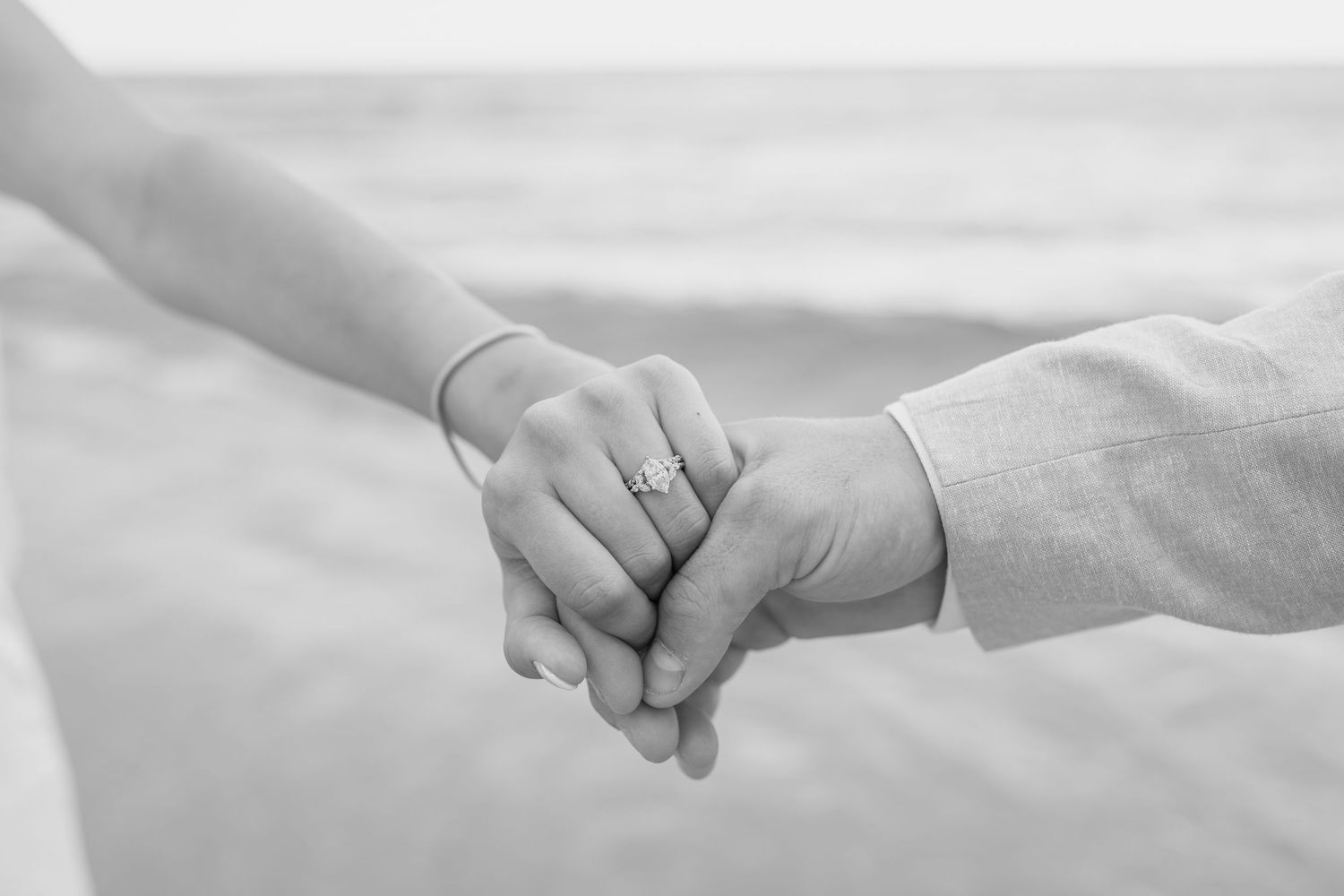 Couple holding hands tenderly with engagement ring visible on sandy beach.