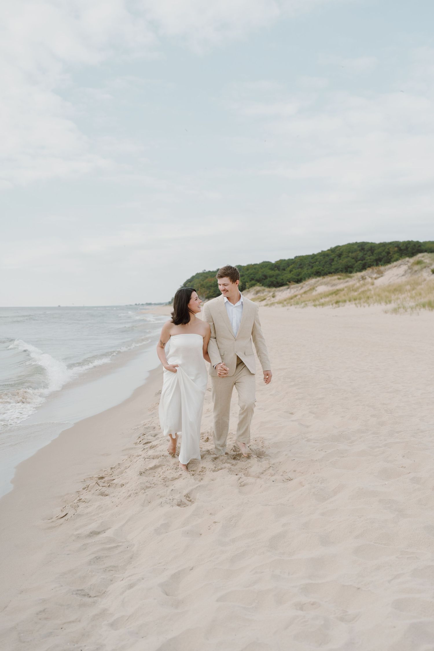 A couple in light summer attire playfully walks along a sandy beach.