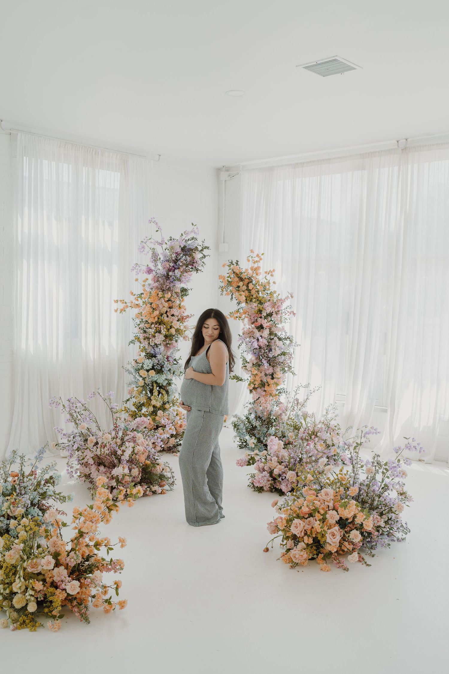 Ethereal maternity portrait with pastel floral installations in white studio setting.