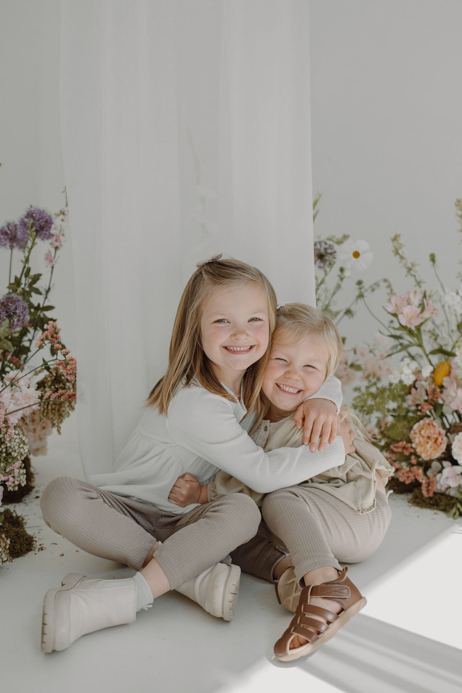 Joyful studio portrait of siblings hugging amid elegant flower arrangements in soft lighting.