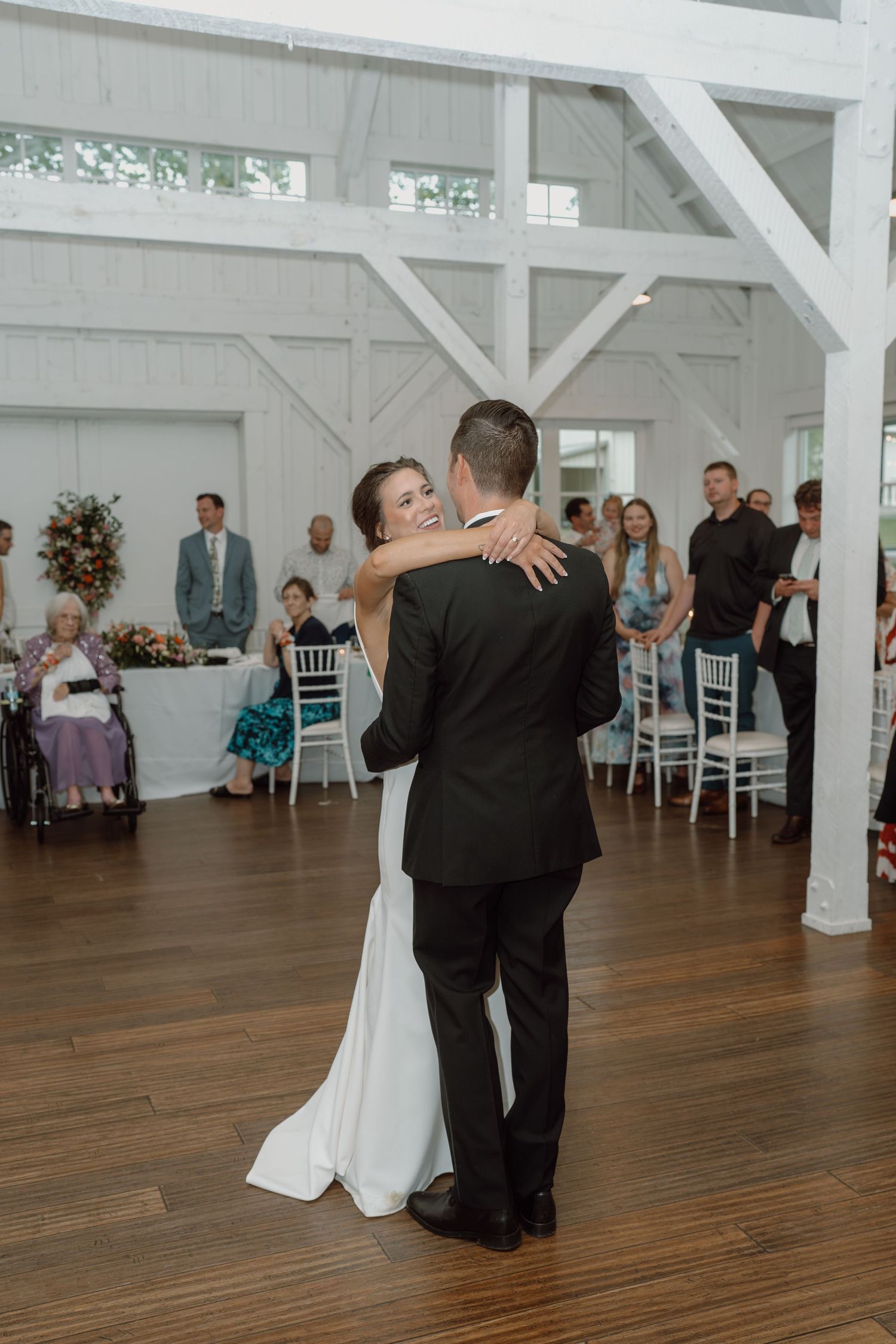 Couple dancing together in an elegant white barn venue while guests look on from their seats.