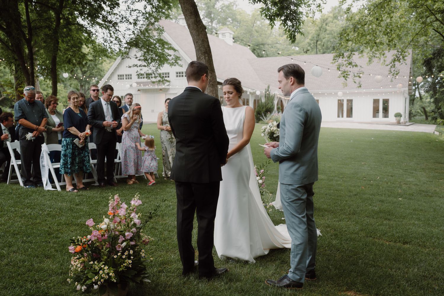 Wedding guests gathered on a lawn for an outdoor ceremony under tall trees.