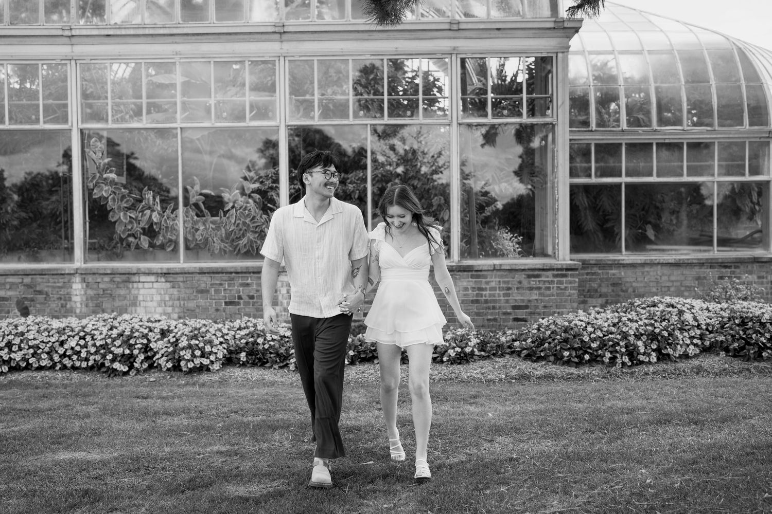 A couple poses together in front of a greenhouse with pink flowers and greenery in the background.