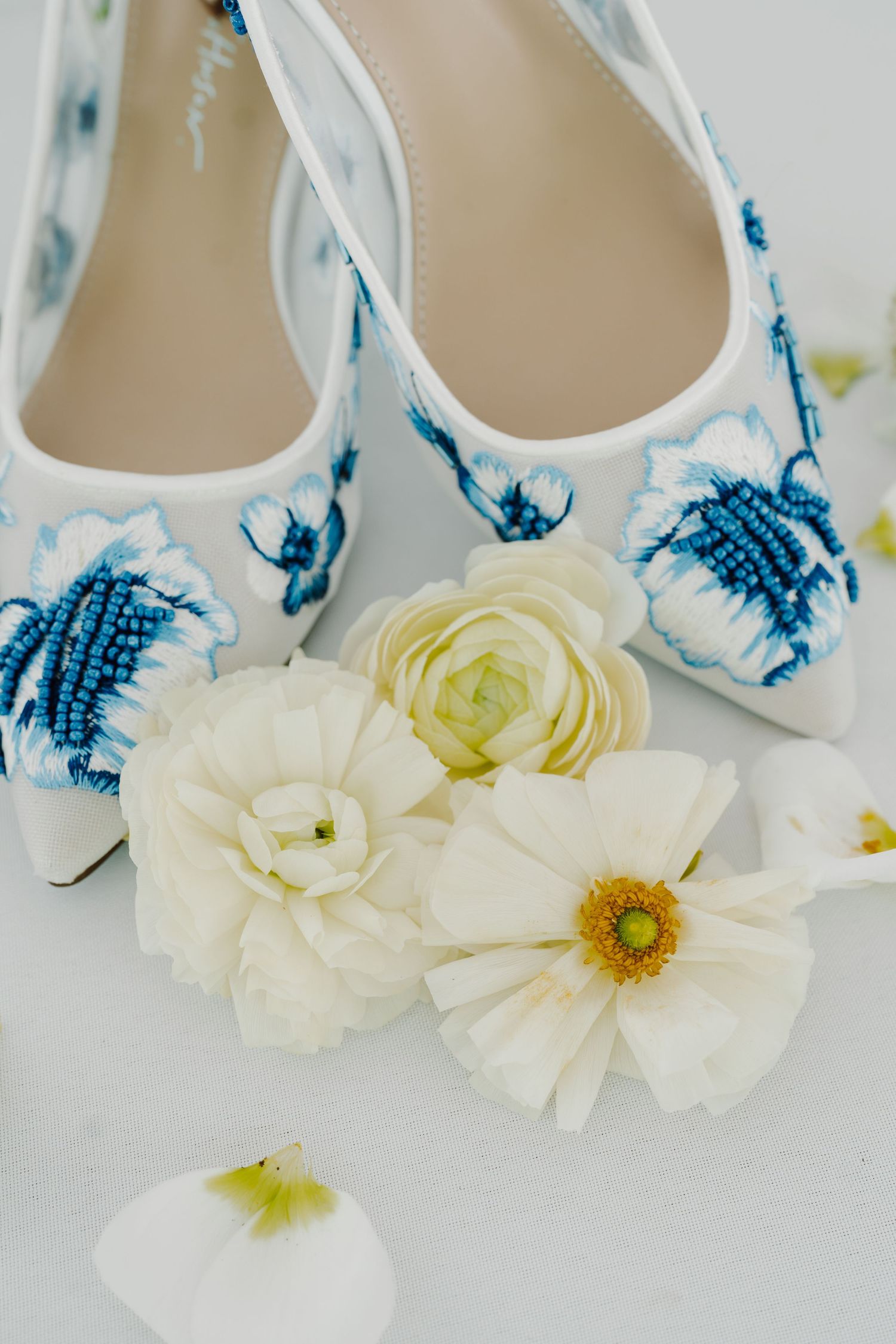 White bridal shoes with blue floral details displayed with white flowers and petals.