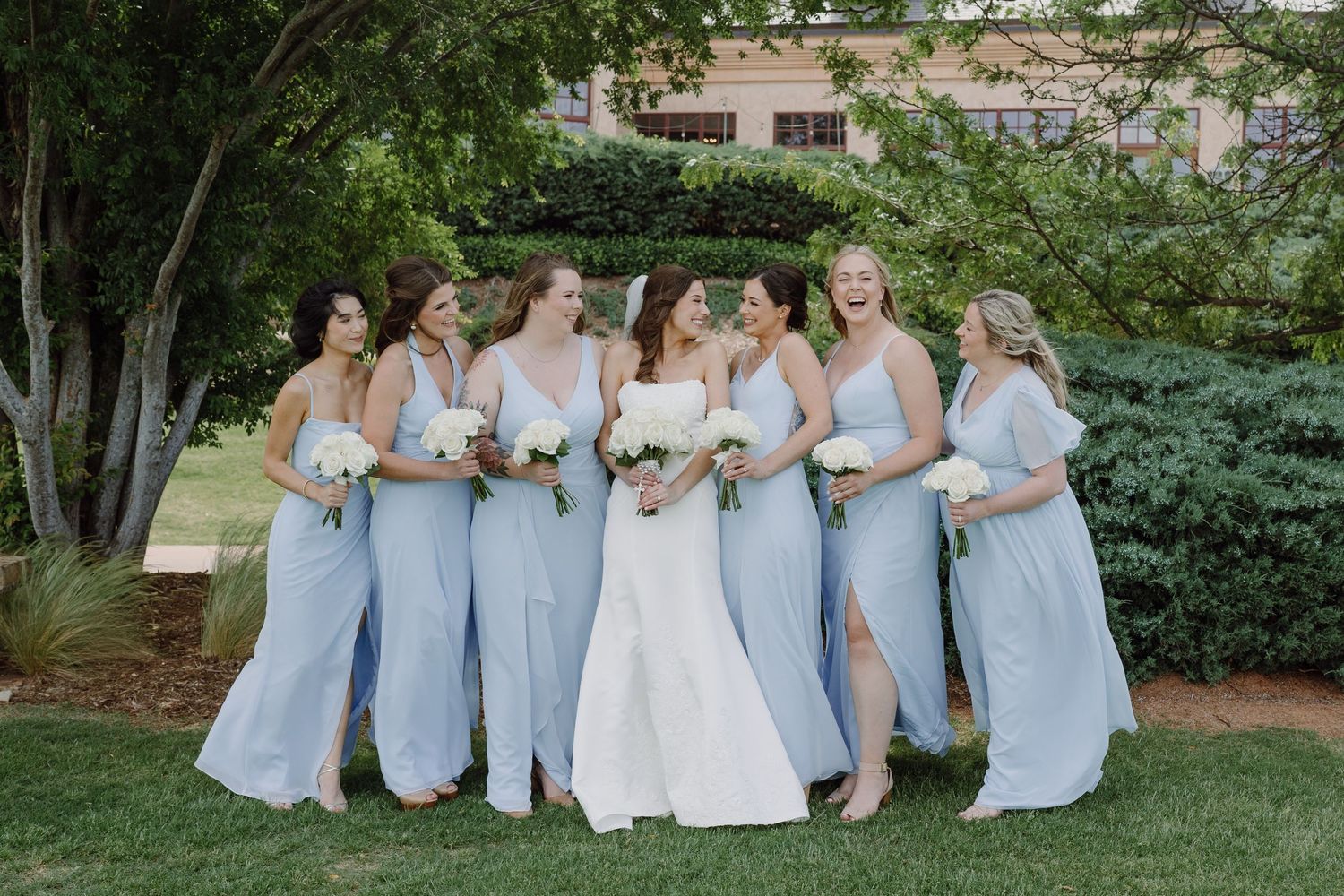A wedding party in light blue bridesmaid dresses and white bridal gown pose together in an outdoor garden setting.