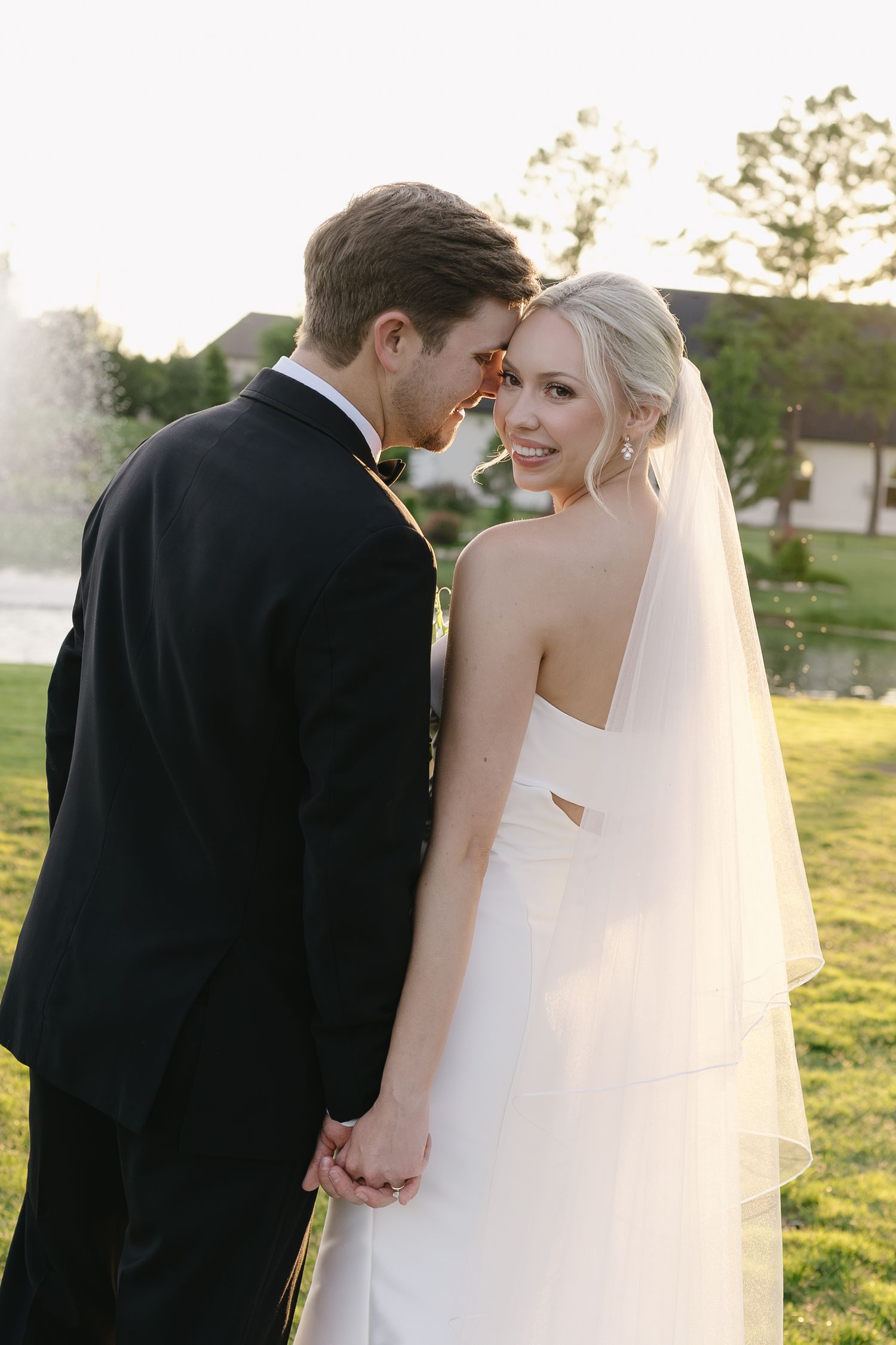 A bride in a white strapless gown and veil holds hands with a groom in a black suit outdoors on a sunny day.