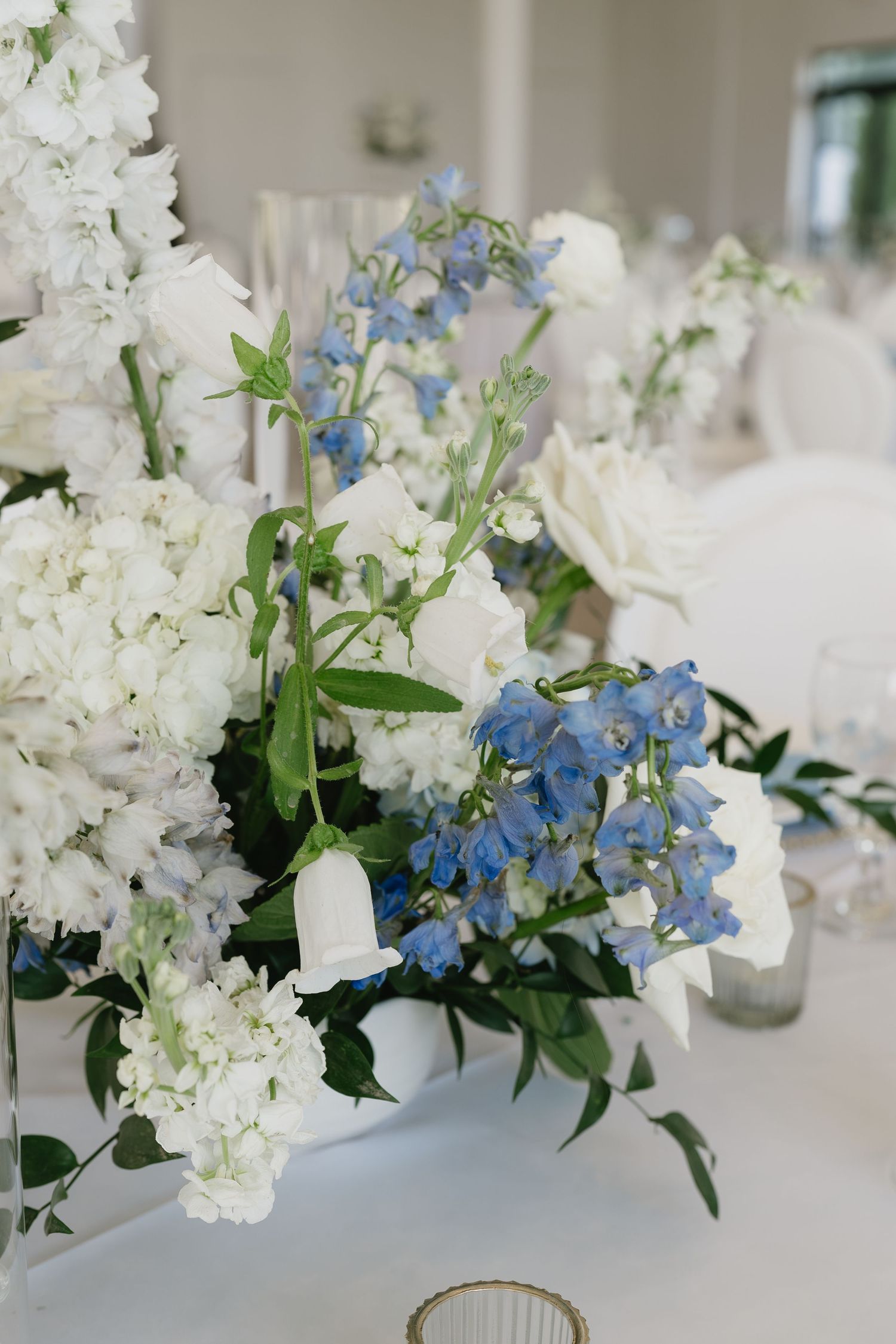 Elegant wedding centerpiece with white hydrangeas, roses, and blue delphiniums arranged in a white vase on a reception table.
