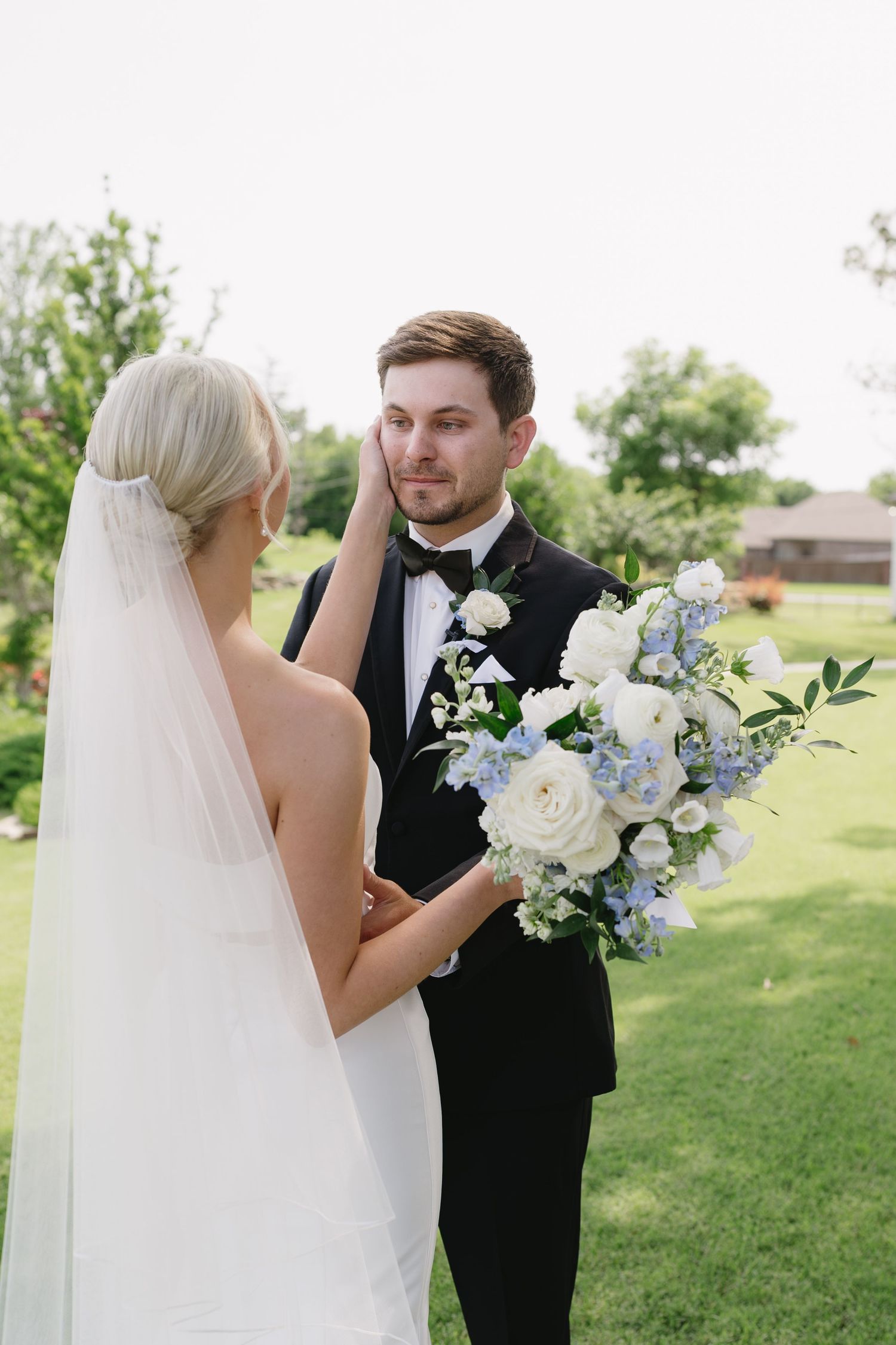 Bride in white veil holds blue and white bouquet while touching groom's face in black tuxedo outdoors.