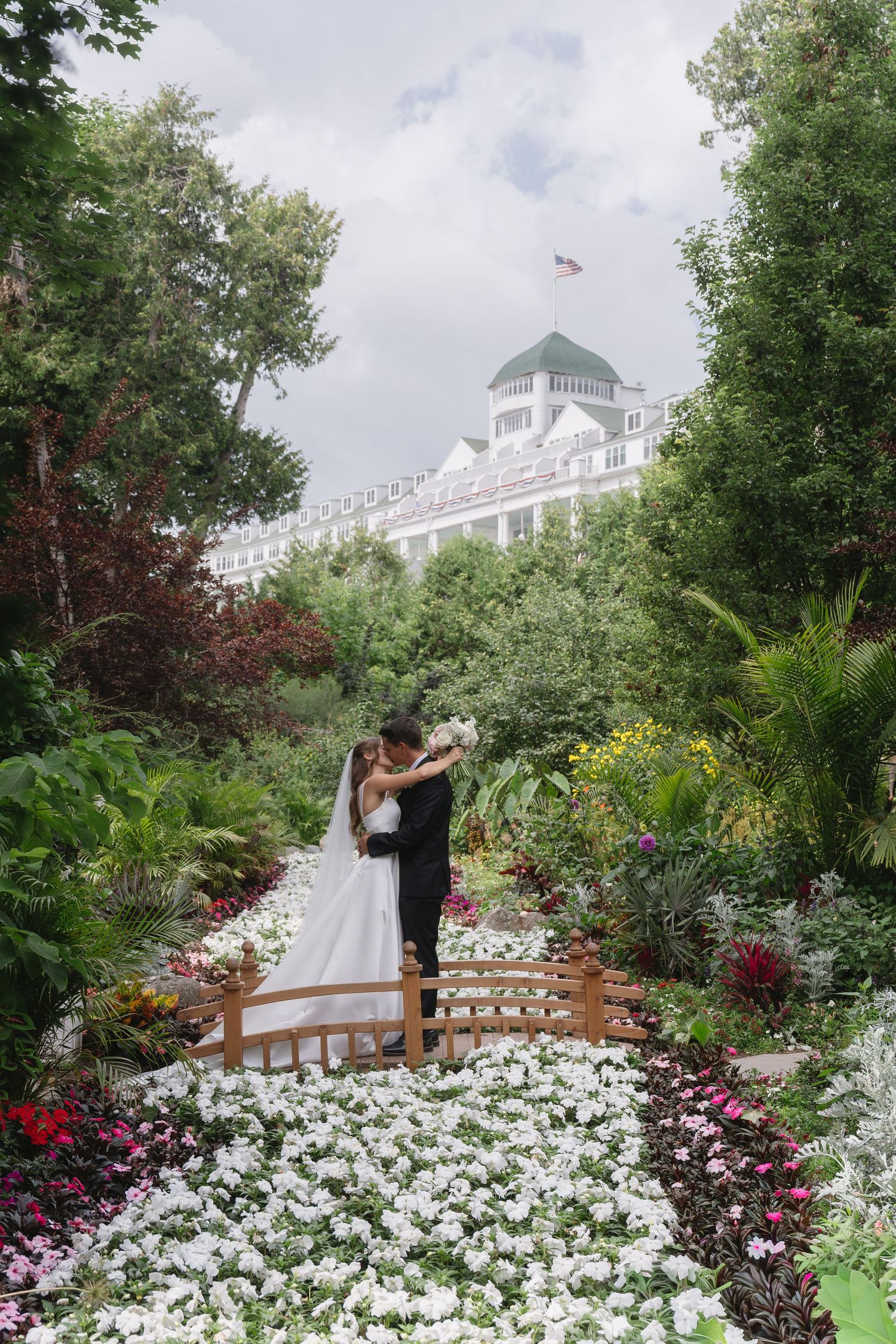 Bride and groom embracing on a wooden bridge surrounded by colorful garden flowers with a grand white hotel in the background.