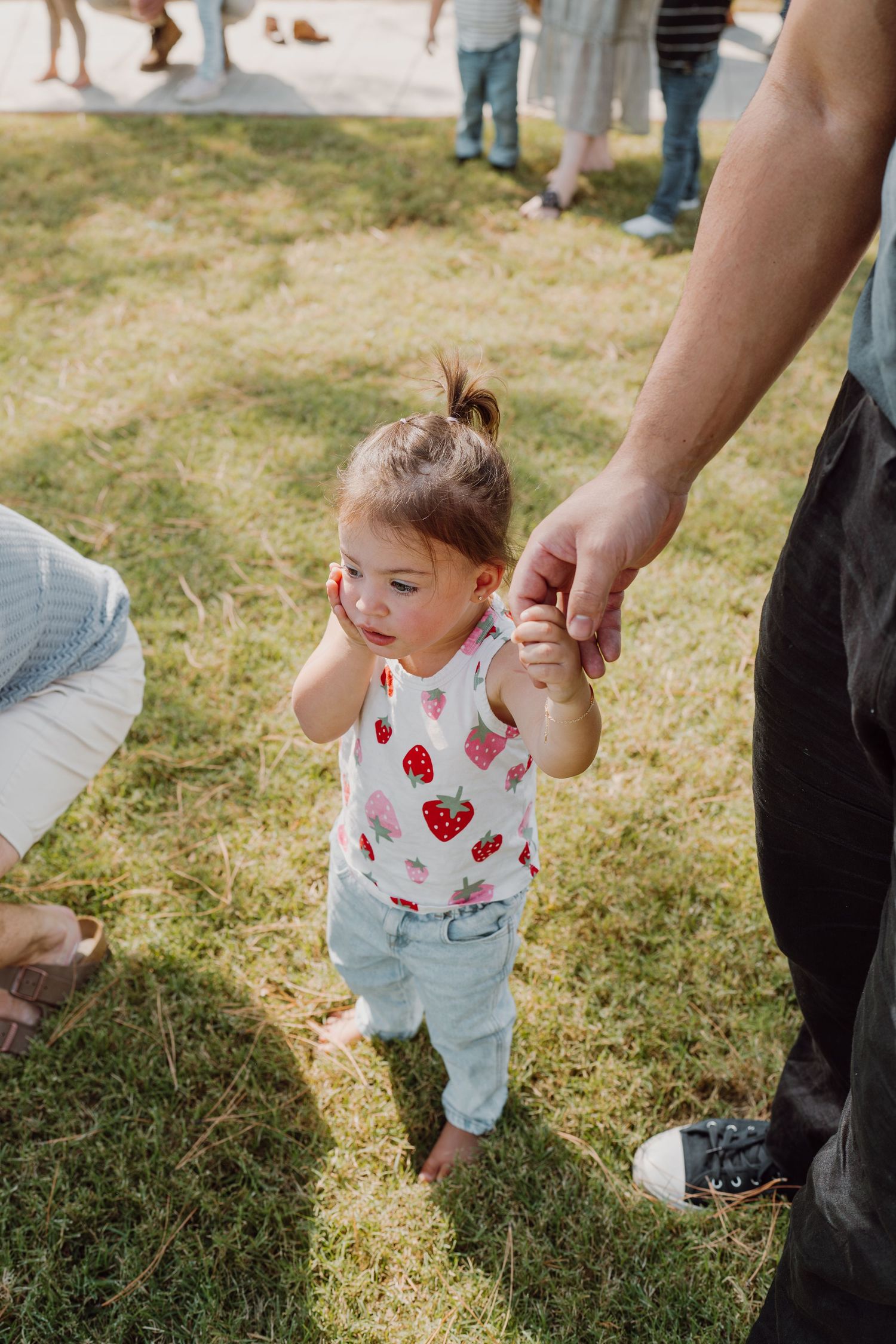 Small child in overalls takes steps on grass while being supported by adults.