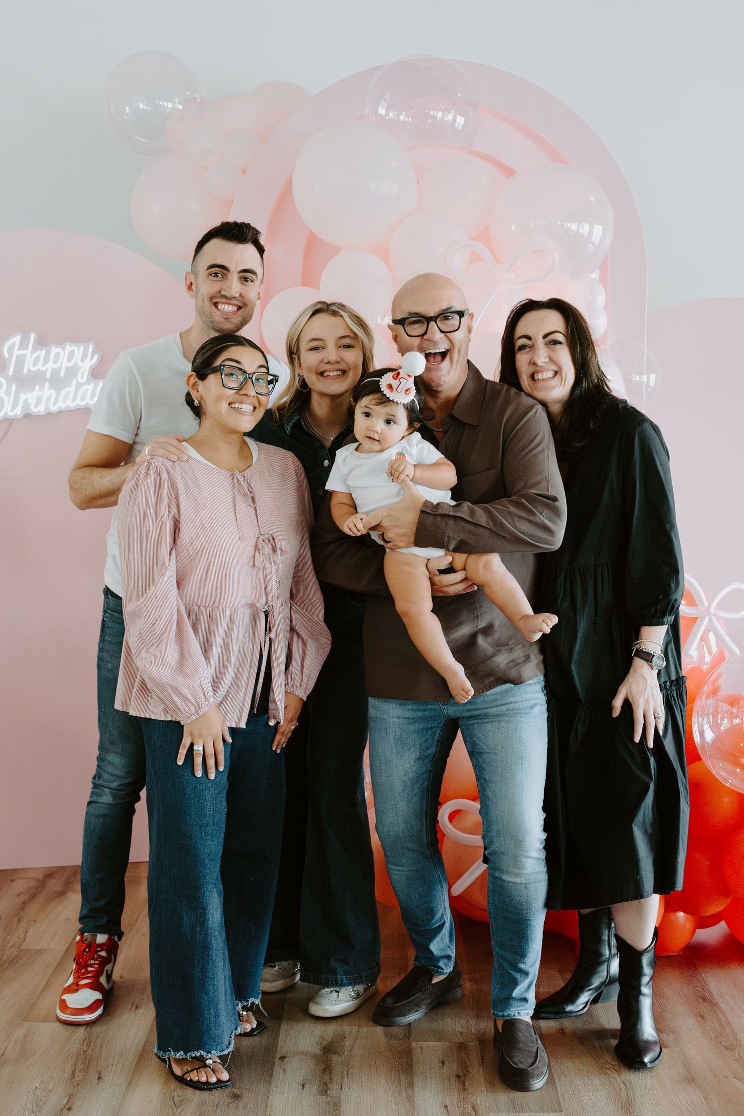 A small group gathers for a photo with a baby in front of a pink and orange balloon display.