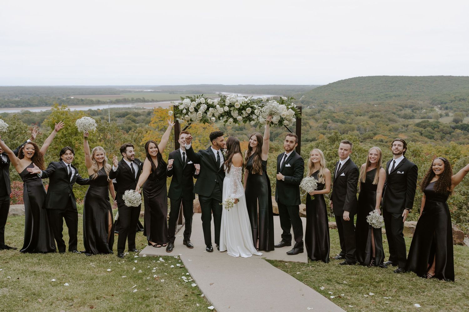 Wedding party in formal attire gathered on a scenic hilltop for group photos.