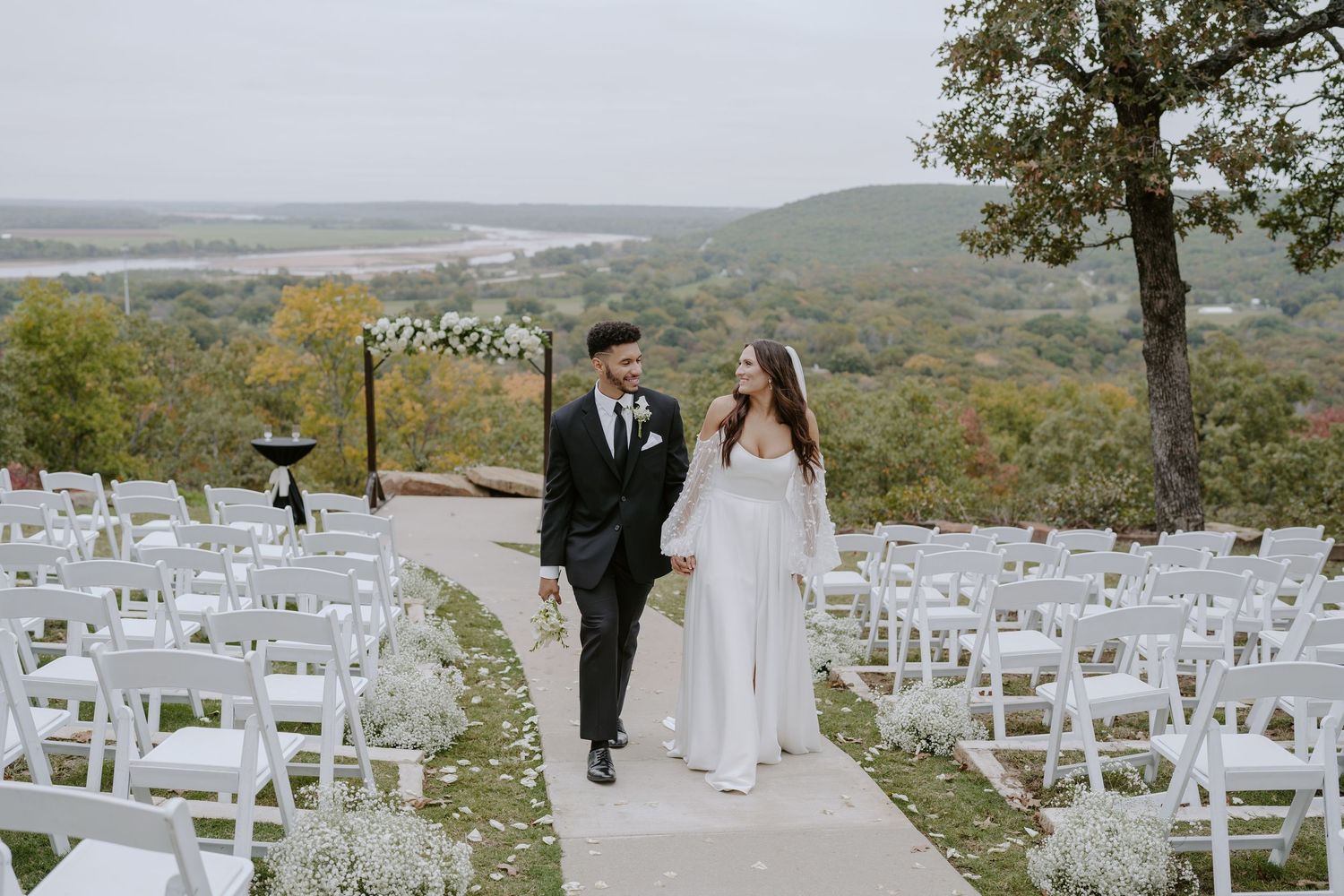 A scenic outdoor wedding ceremony setup with white chairs overlooking rolling hills and autumn foliage.