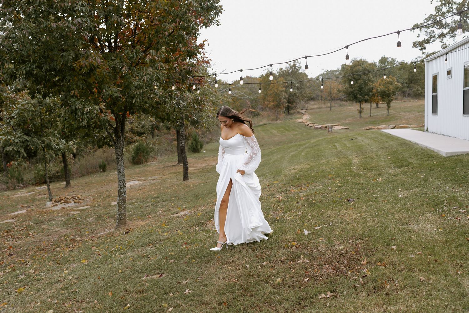 Elegant white wedding dress flows in breeze as person poses on grassy hill near white building.