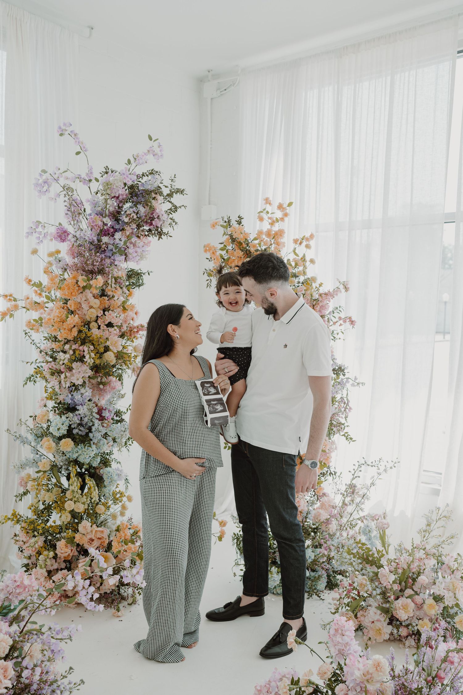 A family poses together in front of a floral arch backdrop while holding ultrasound photos.