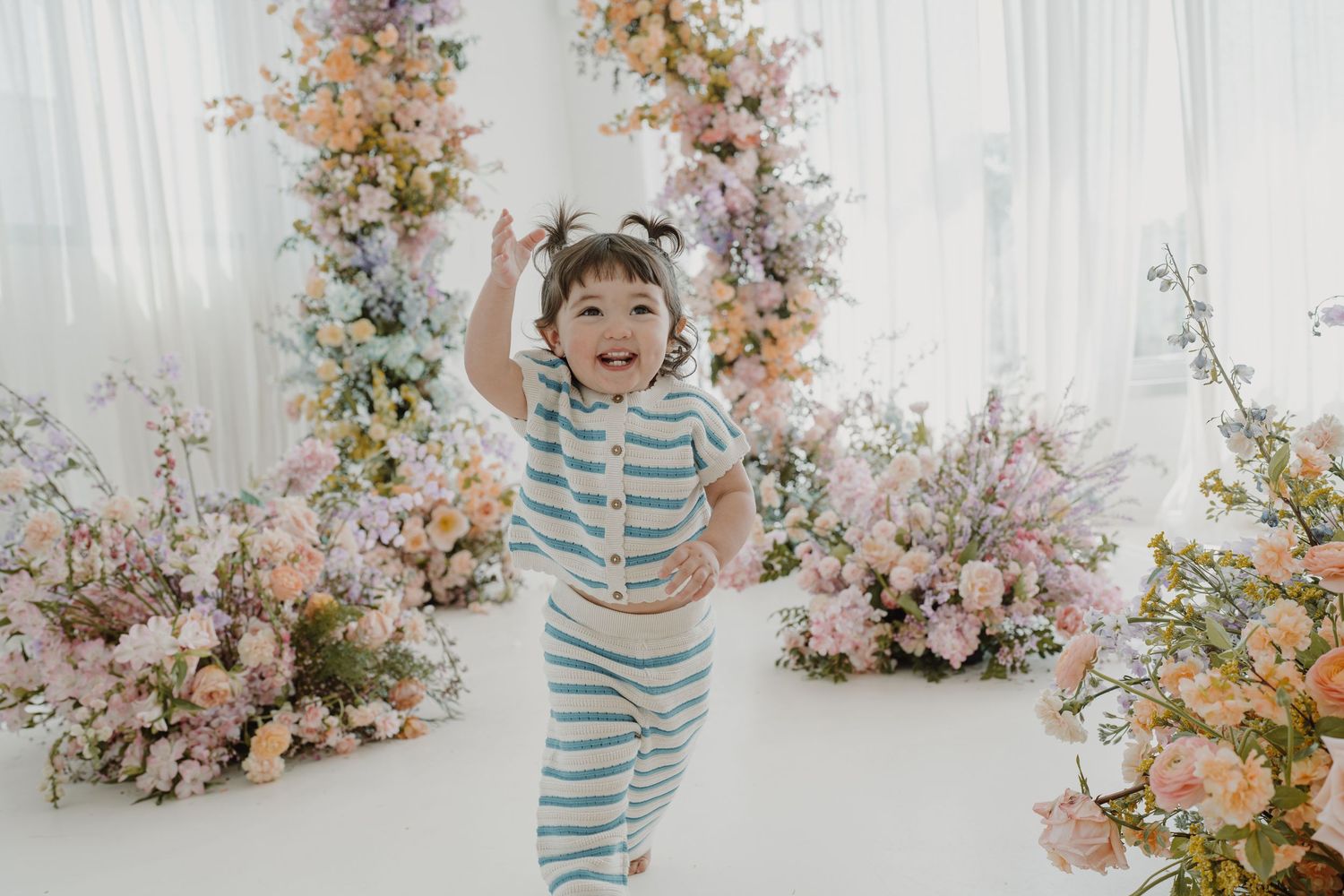 Joyful child in striped outfit jumping with raised arms among pink and peach flowers.