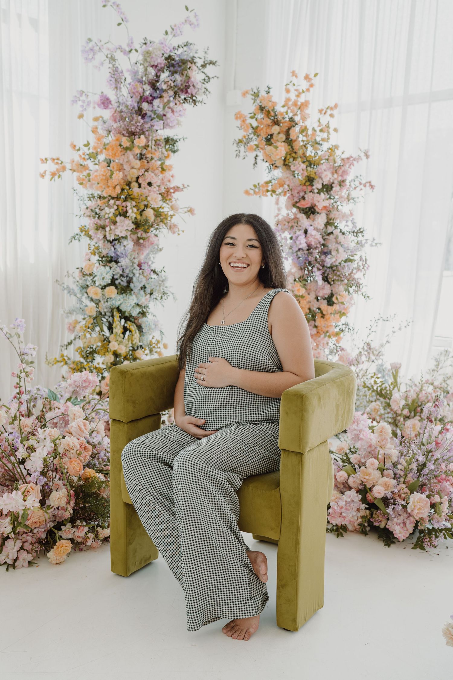 Maternity portrait with elegant floral backdrop and olive green chair in bright studio setting.