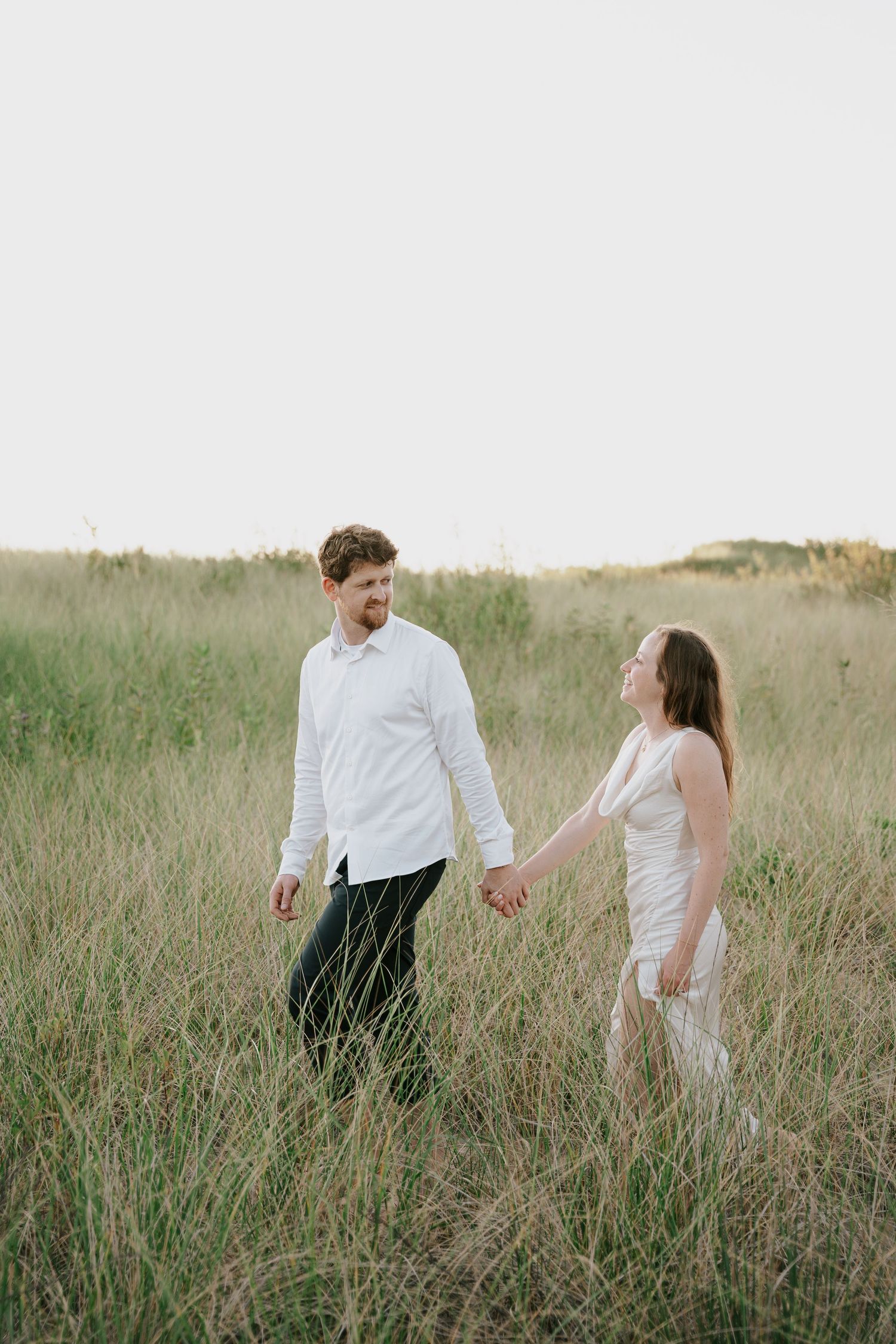 Two people walk through tall grass in a meadow during a romantic photoshoot.